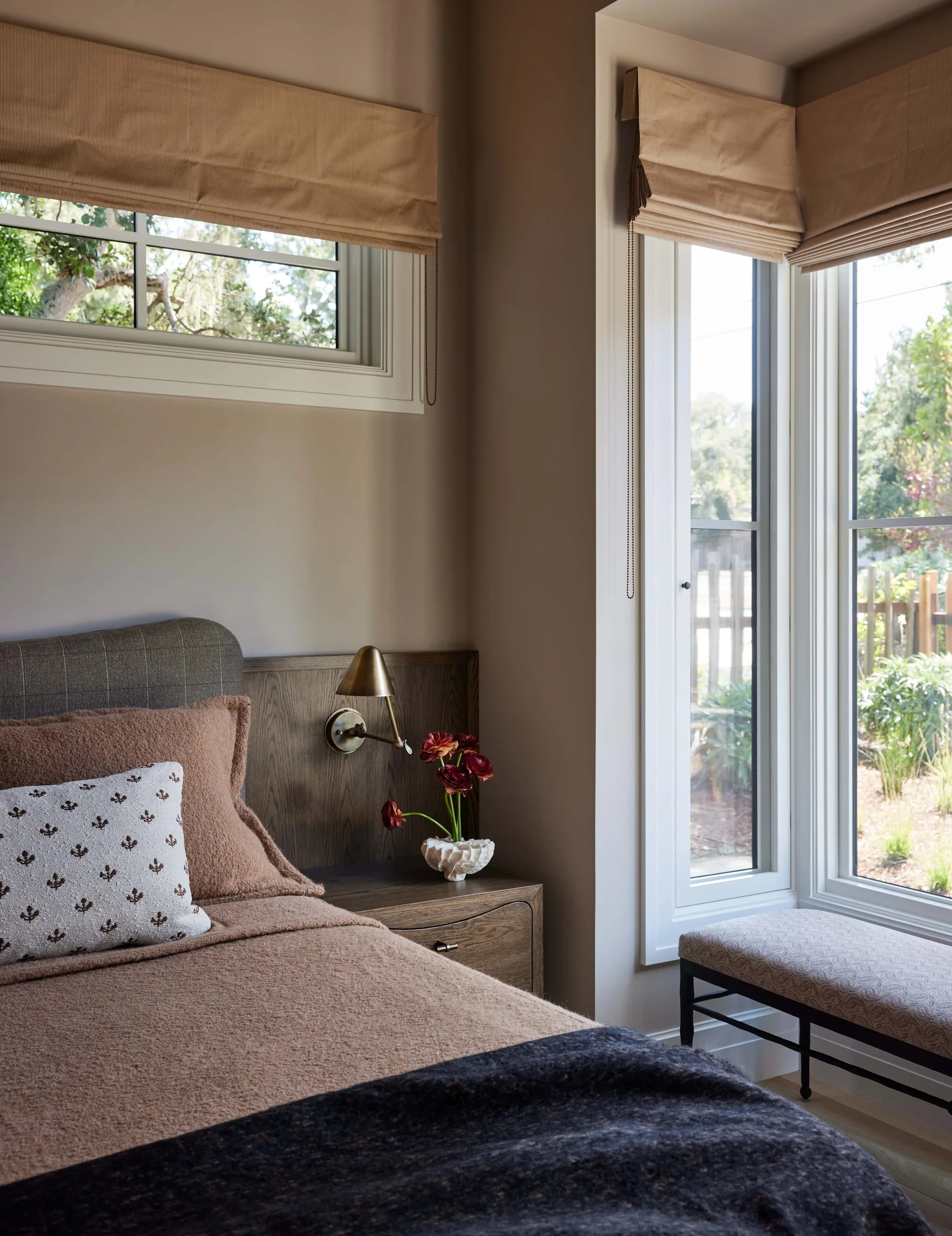 A bedroom corner features a bed with a wooden headboard, a brass wall sconce above a nightstand, and a bay window with tan Roman shades.