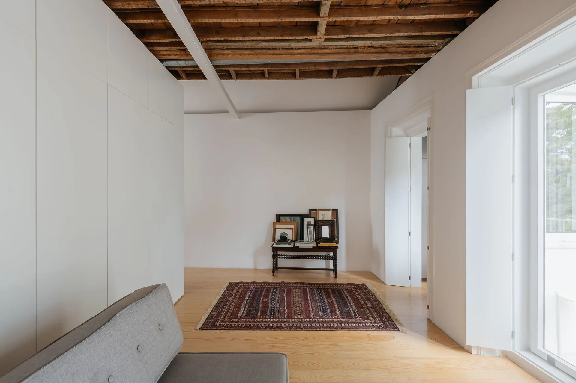 A minimalist living room featuring exposed wooden ceiling joists, clean white walls, a grey sofa, and a patterned red rug on light wood floors.