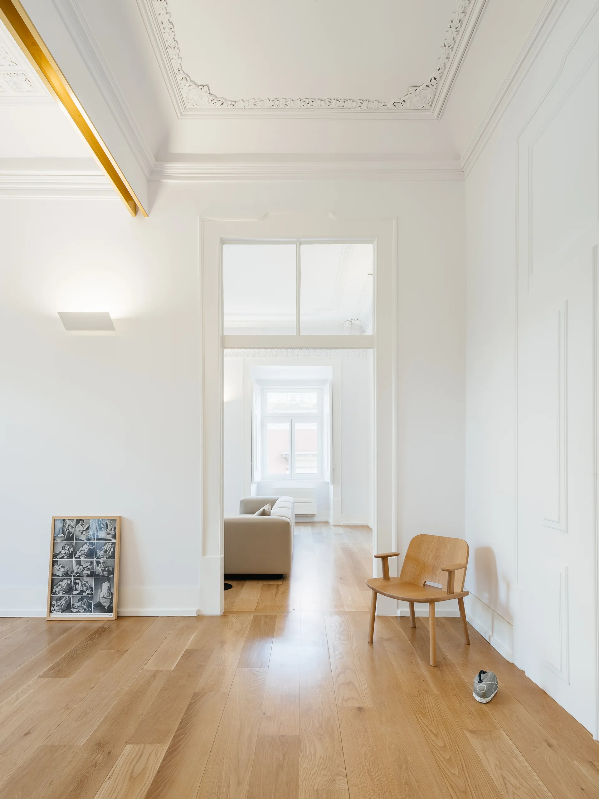 A minimalist room features light wood floors, white paneled walls, a single wooden chair, and an ornate white ceiling with decorative plaster molding.