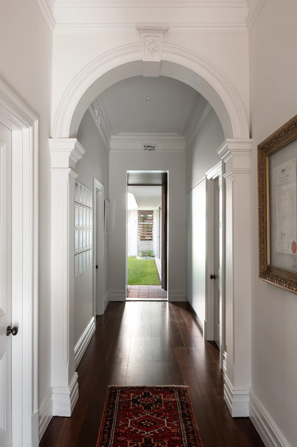 A bright white residential hallway featuring dark hardwood flooring, detailed crown molding, a large central archway, and a view of an outdoor green garden.