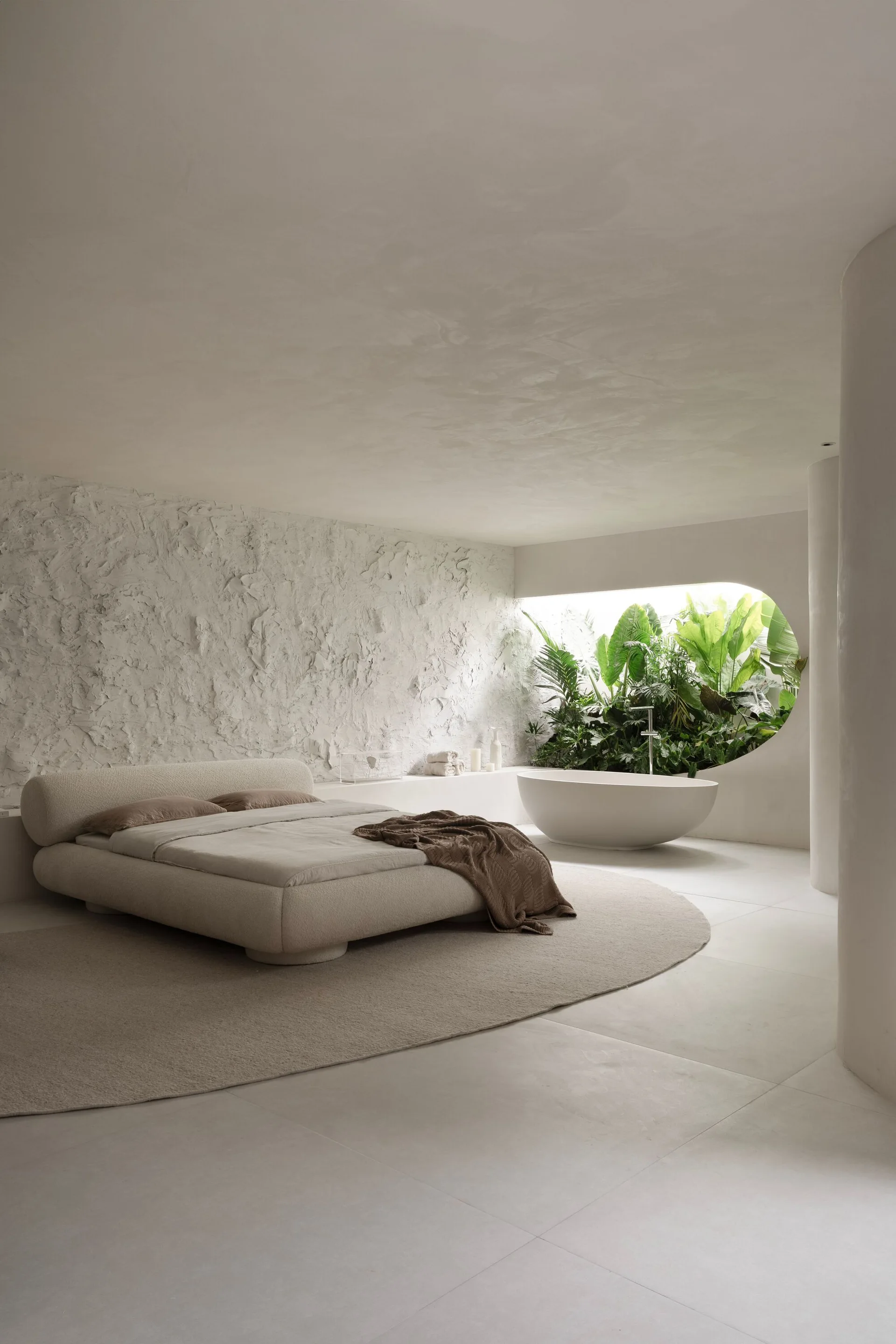 An off-white bedroom features a low platform bed, a textured plaster wall, and a freestanding bathtub in front of an oval window filled with green foliage.