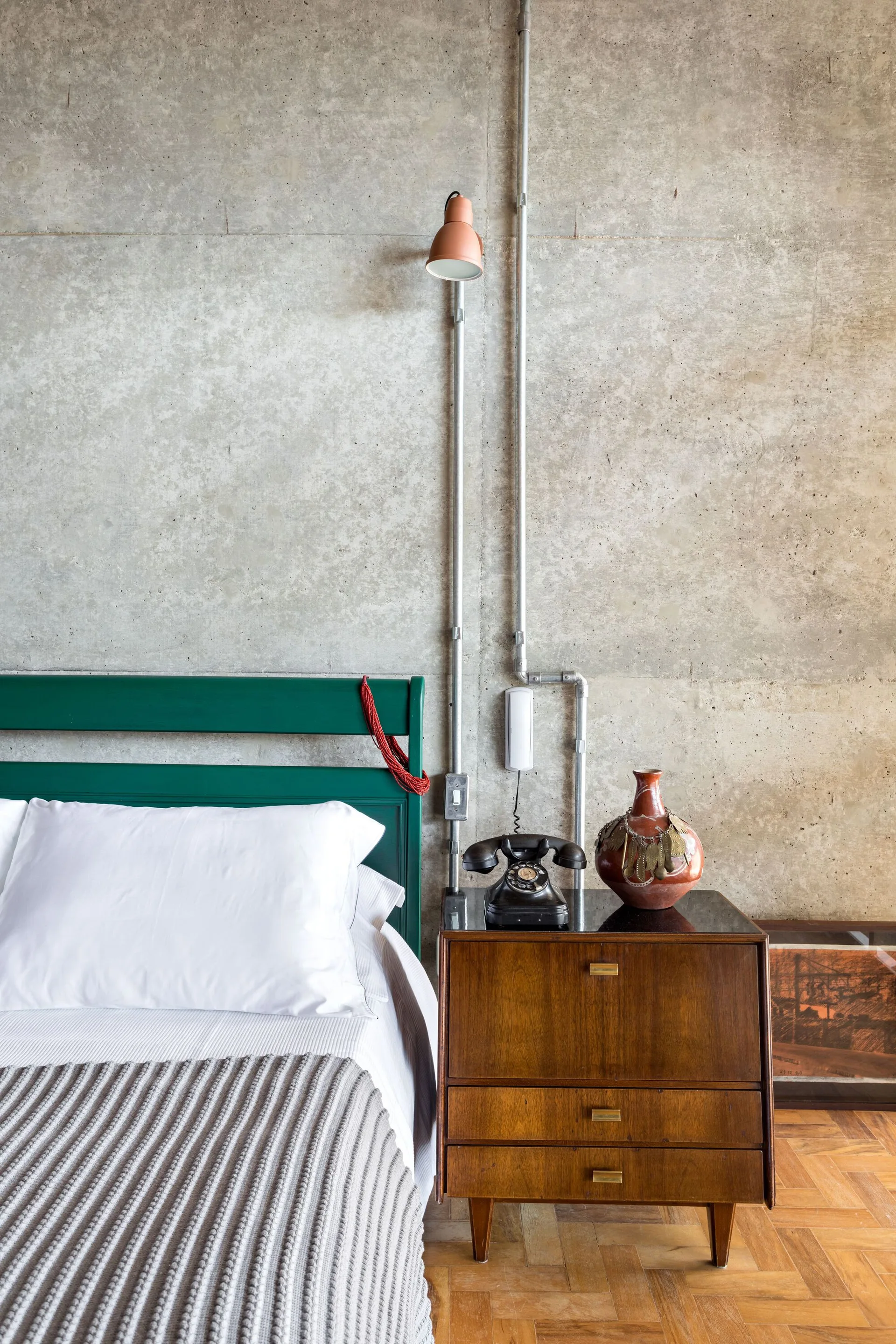 A bedroom features an exposed concrete wall with metal conduits, a green wooden headboard, and a mid-century wooden nightstand.