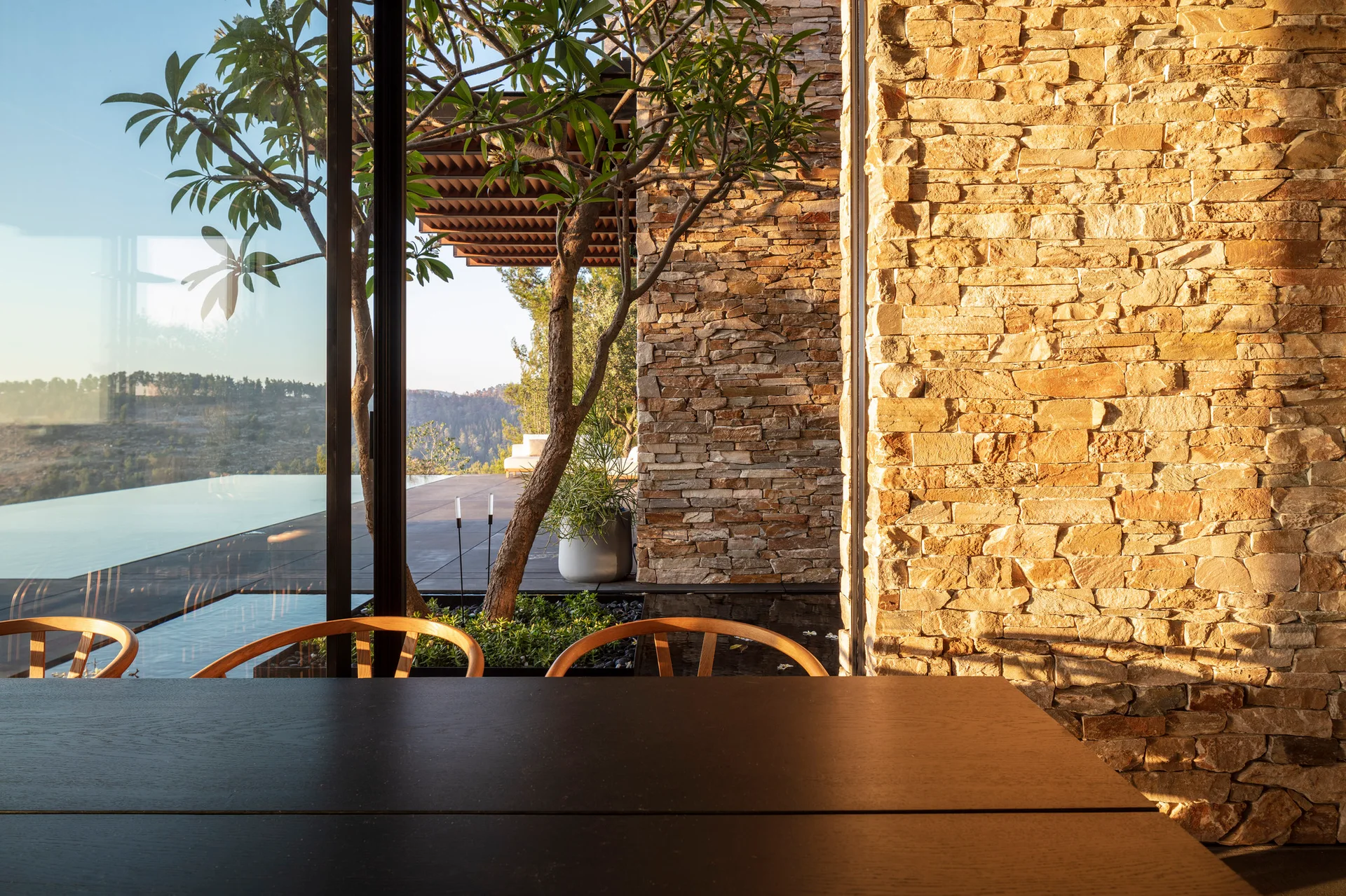 A modern dining room features a dark wood table and chairs beside a floor-to-ceiling glass wall overlooking a terrace pool and a textured stone wall.