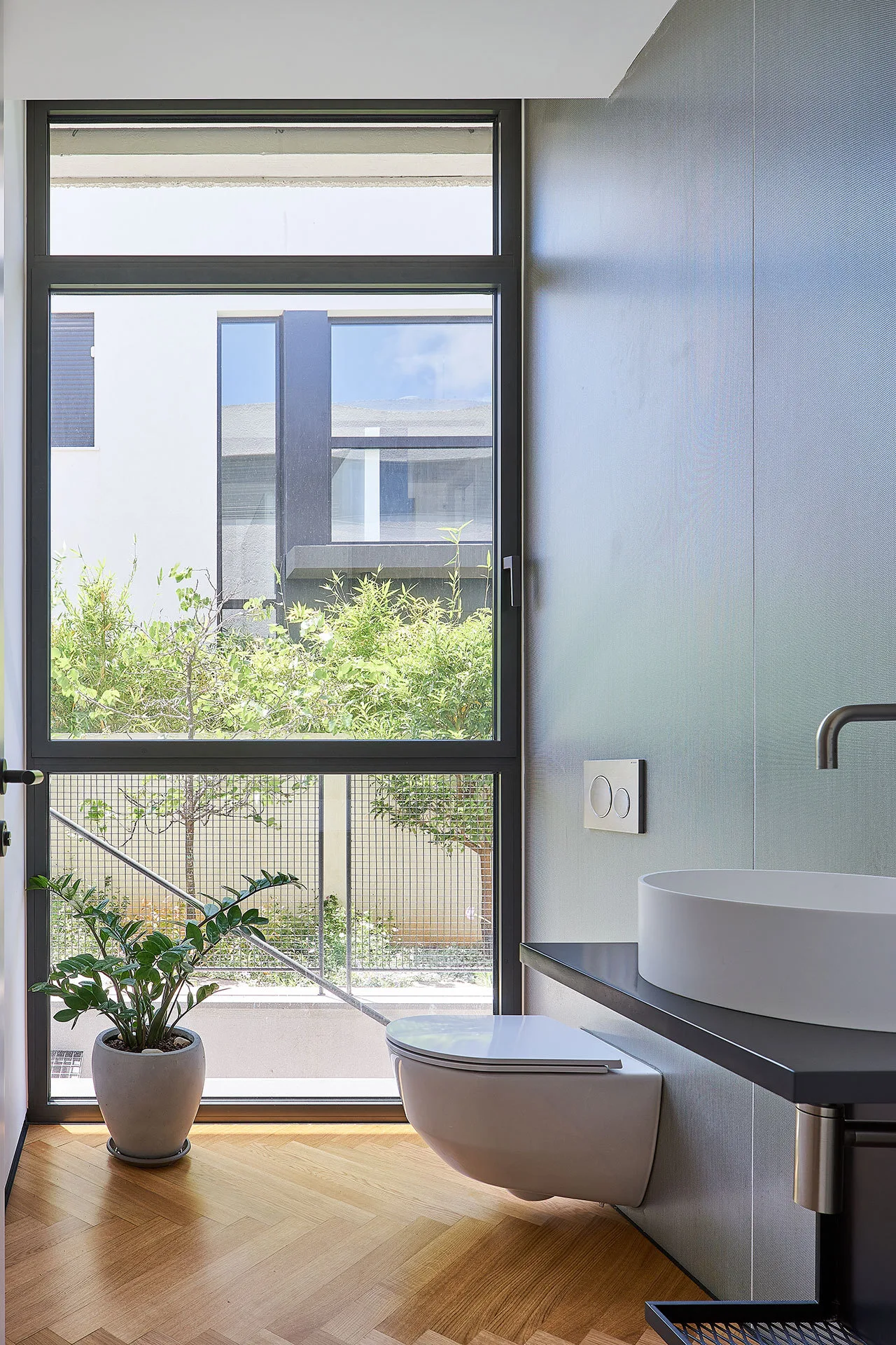 A modern bathroom features a wall-mounted toilet, a white vessel sink on a dark counter, herringbone wood flooring, and a large floor-to-ceiling window.