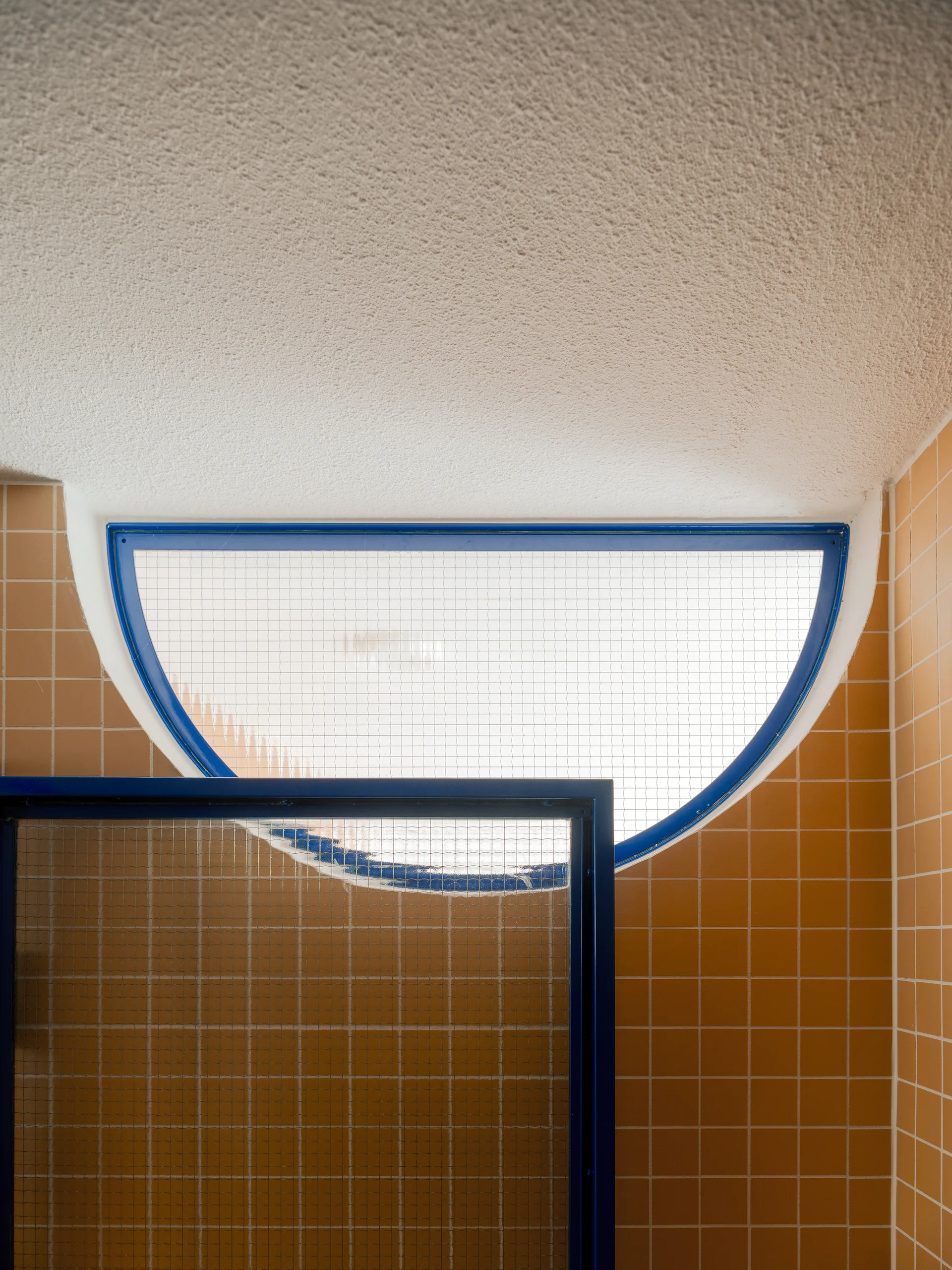A semicircular window with a bright blue metal frame is set into a wall of glossy orange square tiles above a matching blue-framed glass partition.