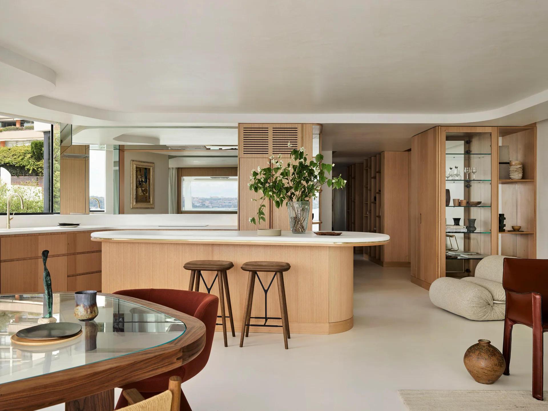 A light wood kitchen island with a curved white top sits beside matching oak cabinetry, a glass dining table, and bar stools.