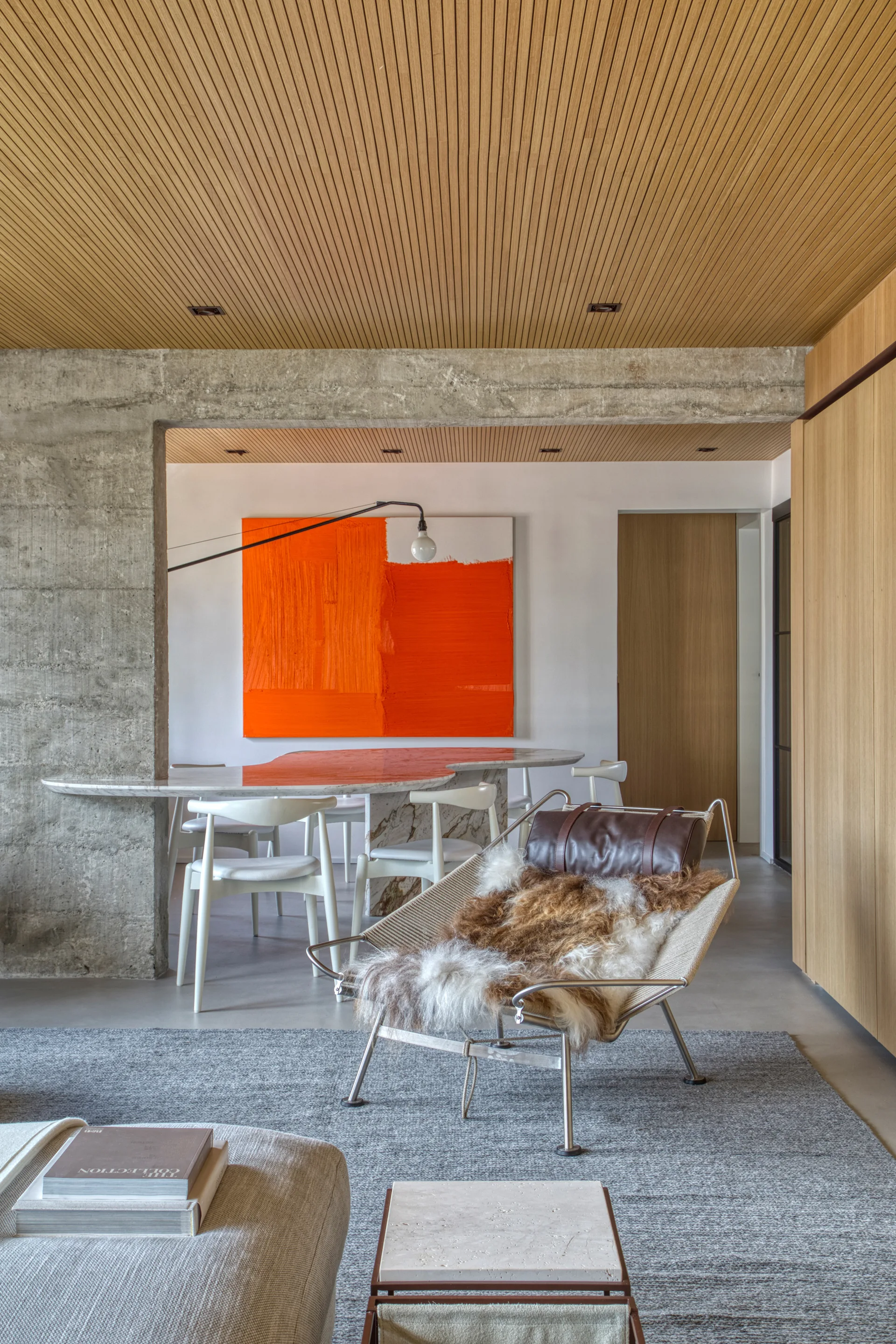 A modern open-concept living area features a slatted wood ceiling, a raw concrete wall, a lounge chair with a fur throw, and large orange art.