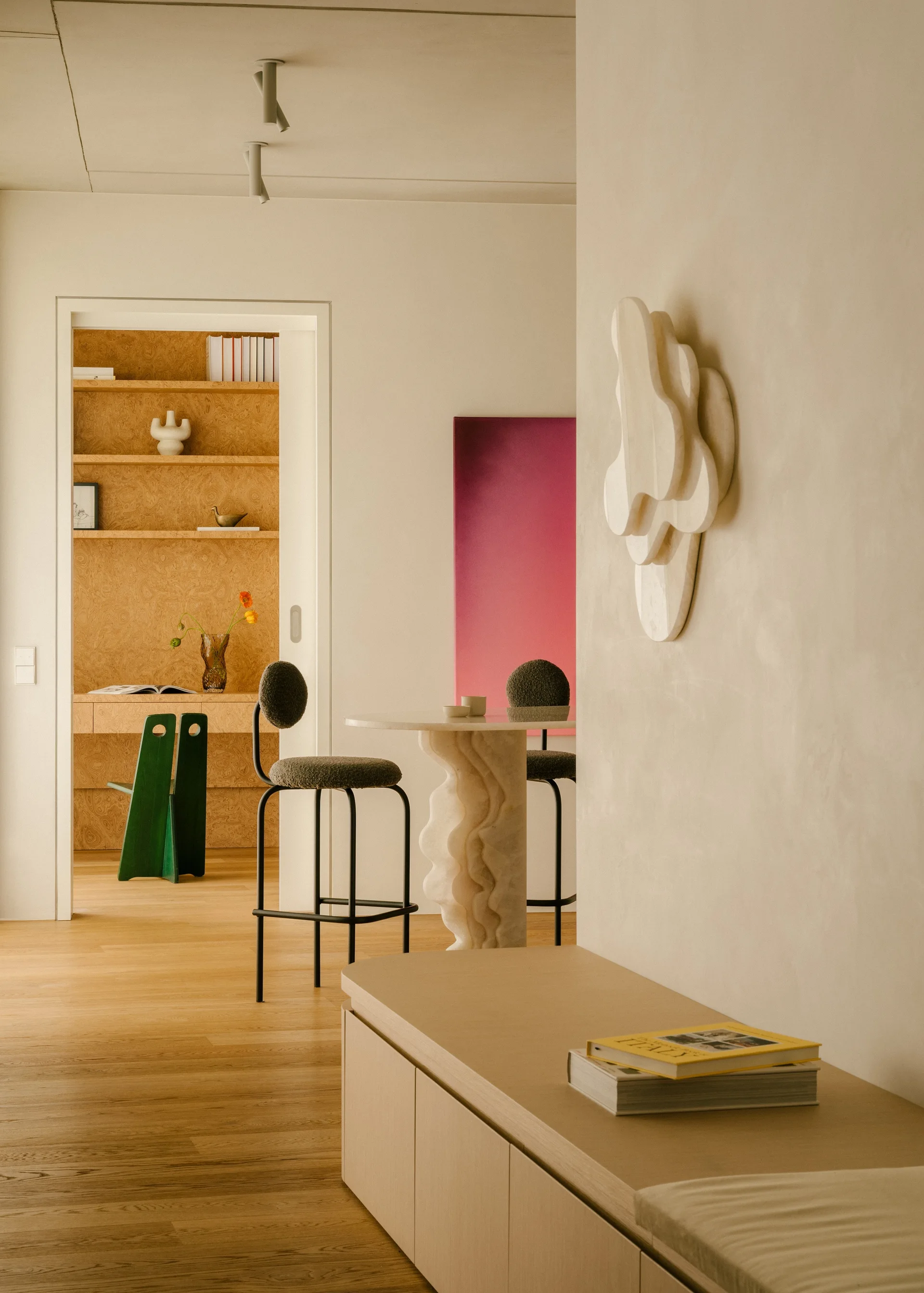 A sculptural stone-base bistro table and two bar stools stand near an open doorway leading to a wood-paneled home office with built-in shelves.
