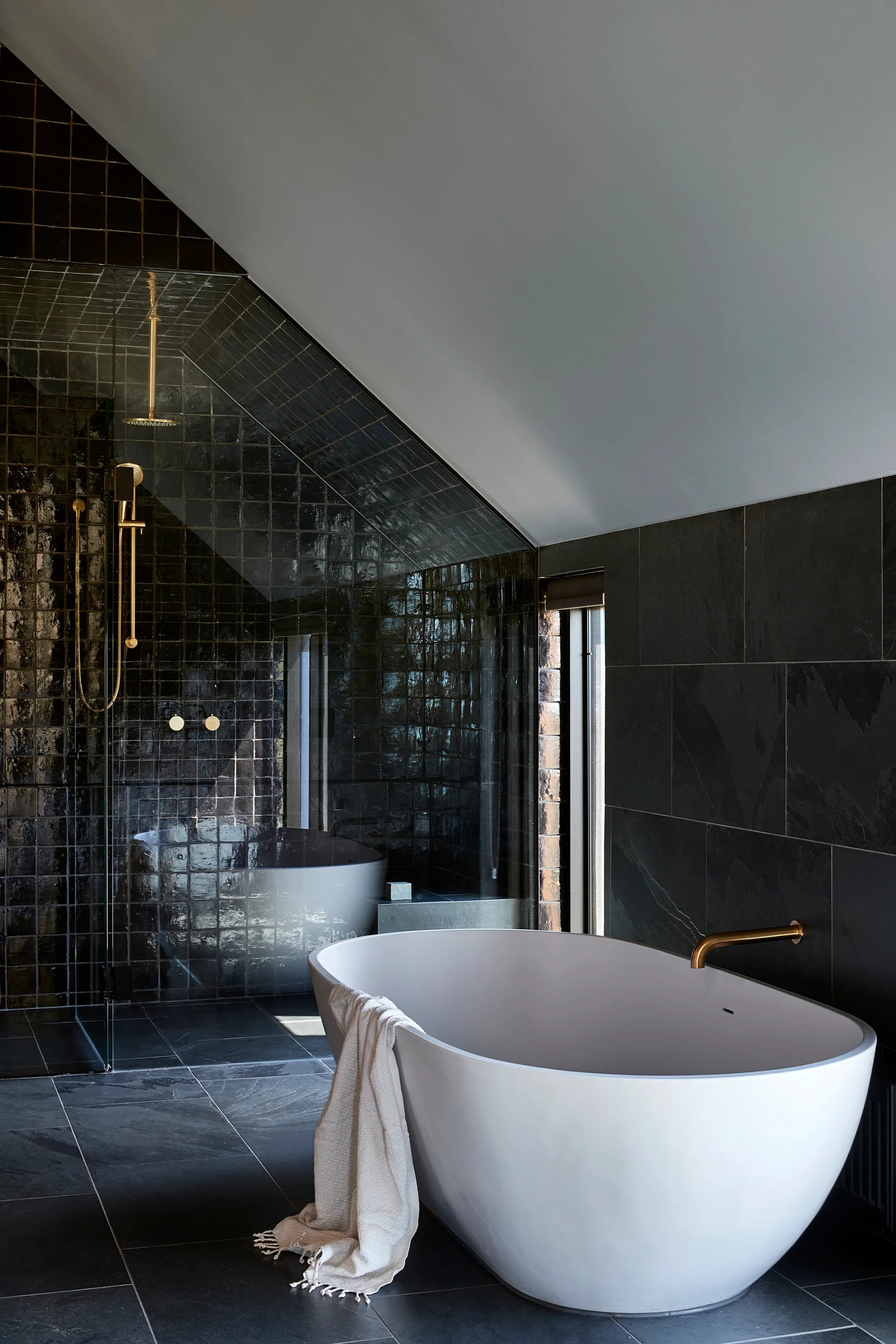 A modern bathroom with a white freestanding bathtub, matte grey floor tiles, and a walk-in shower featuring glossy black wall tiles and brass fixtures.