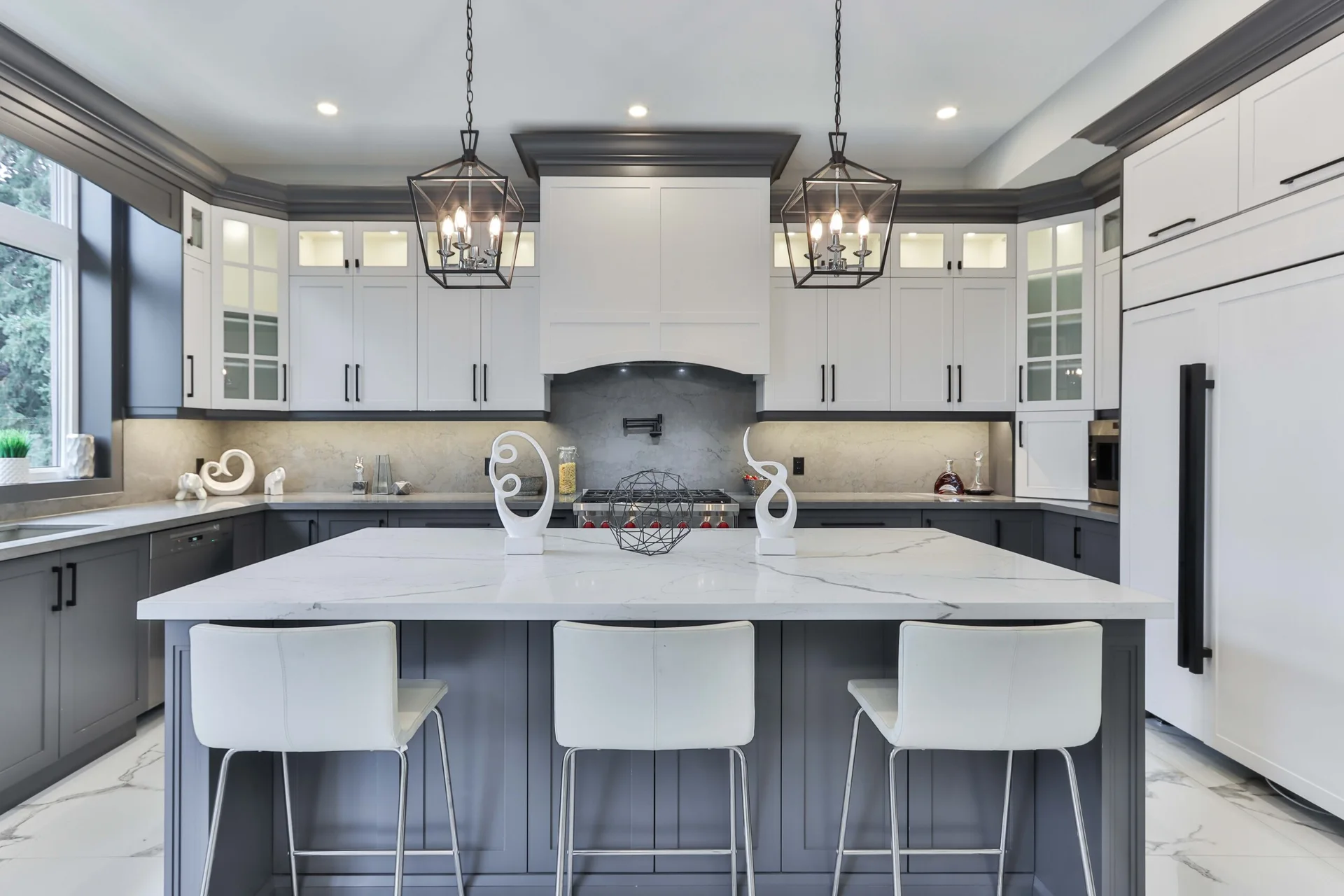 A transitional kitchen features two-toned white and grey cabinets, a marble-topped center island with three bar stools, and geometric pendant lights.