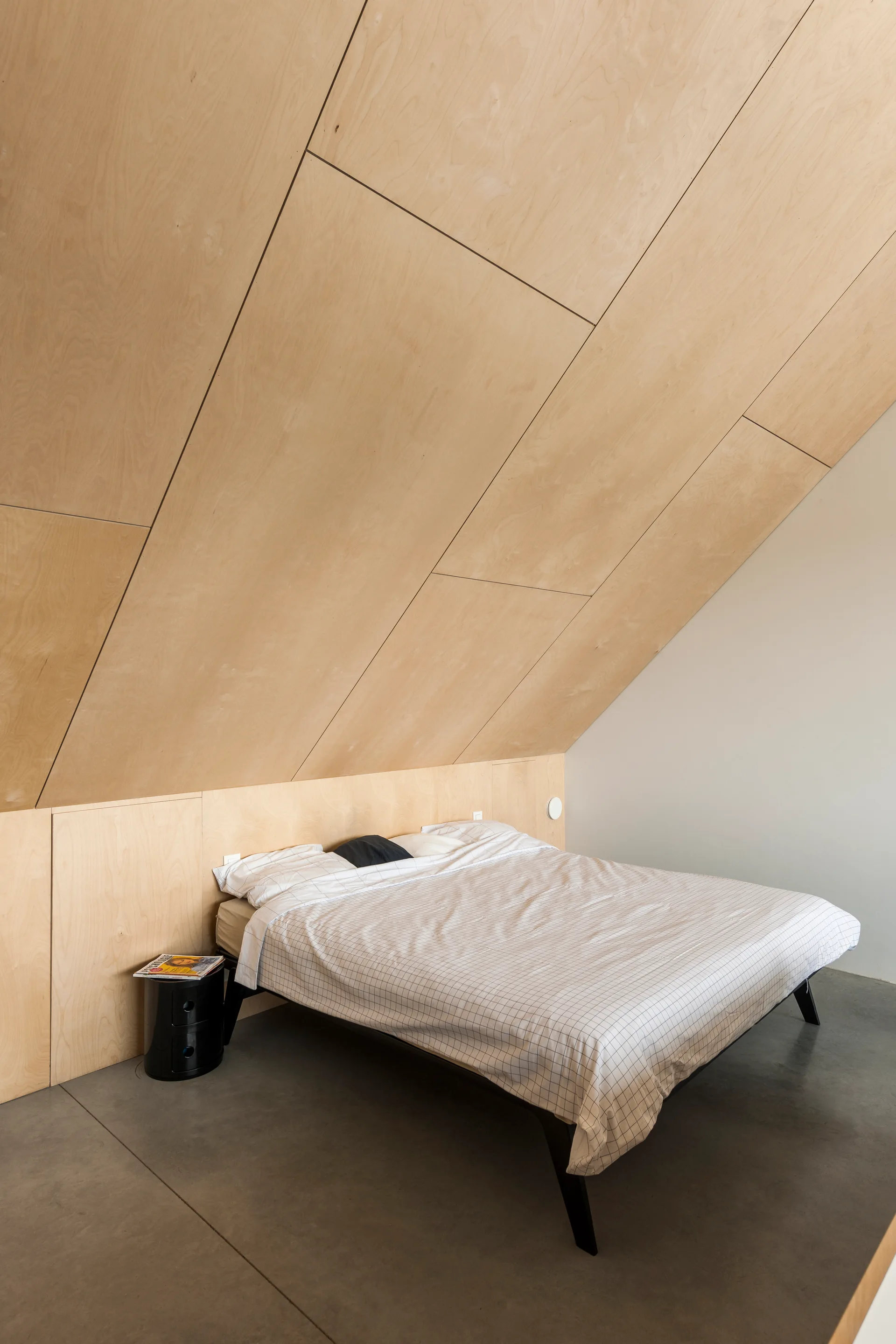 A minimalist bed with grid-patterned bedding sits beneath a sloped plywood ceiling in a bedroom with grey concrete flooring.