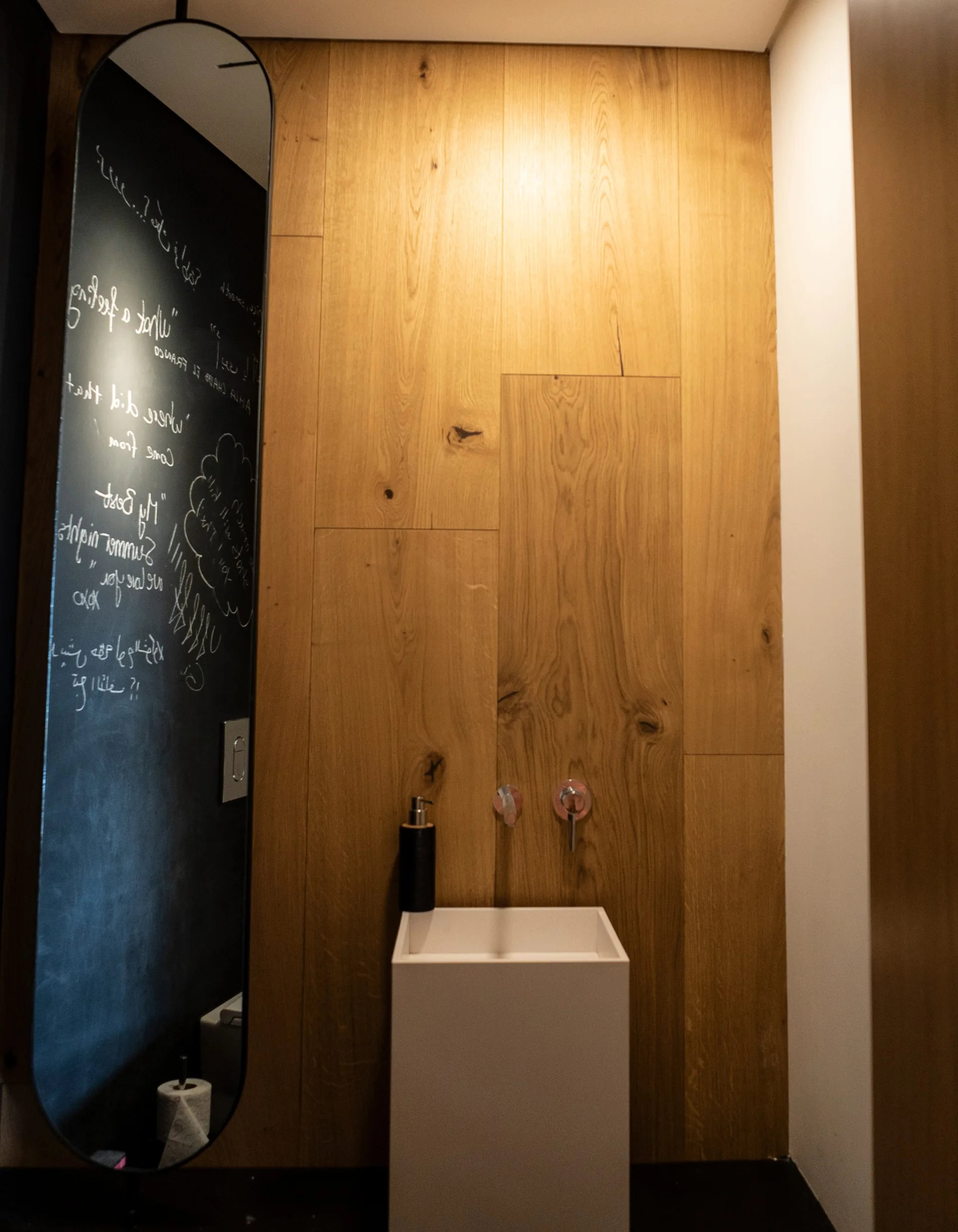 A minimalist powder room features a white square pedestal sink against warm wood wall paneling next to a tall mirror with white script text.