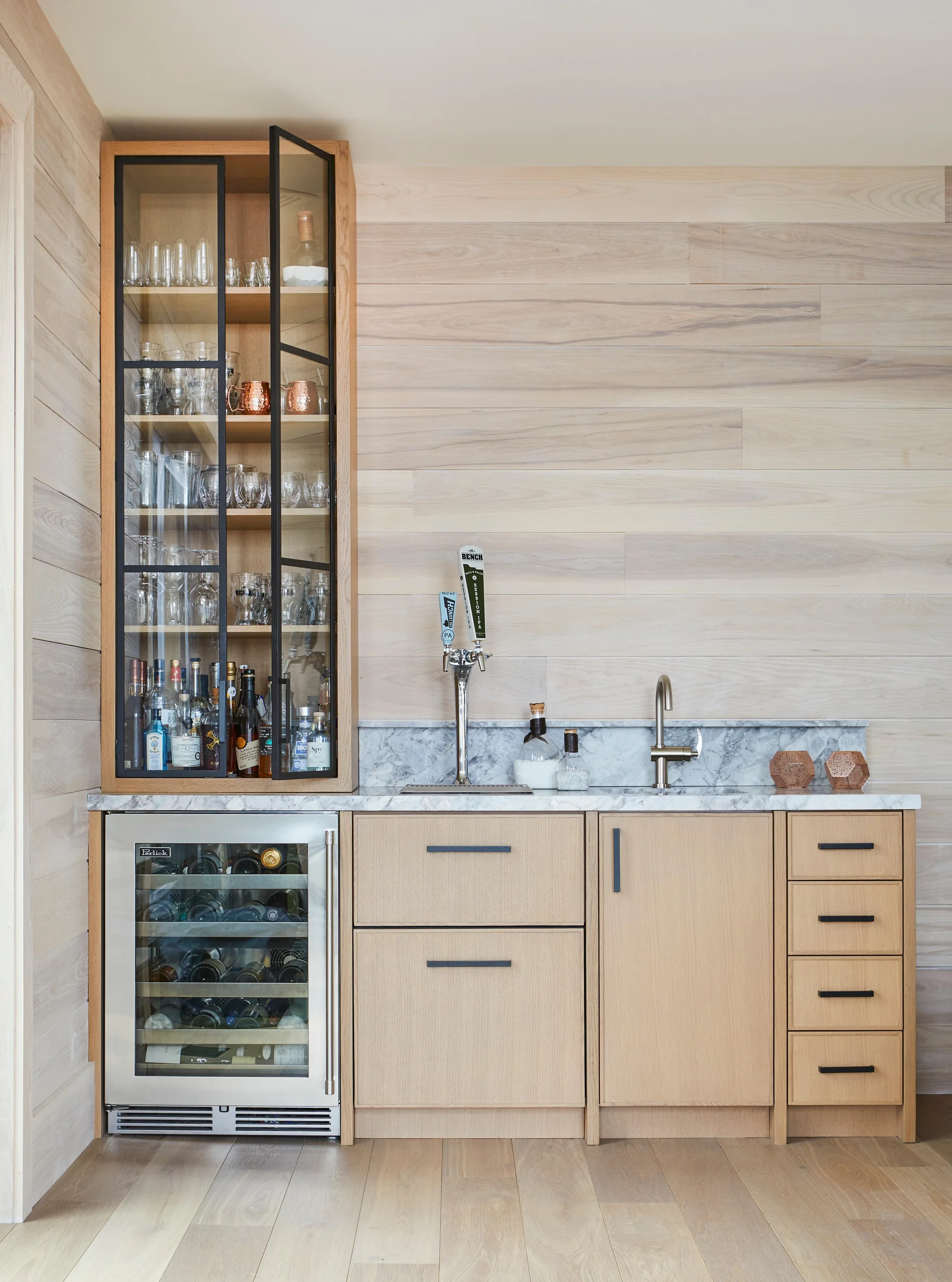 A modern home bar featuring light wood cabinetry, matching horizontal wood wall paneling, a marble countertop, and a glass-front display cabinet.