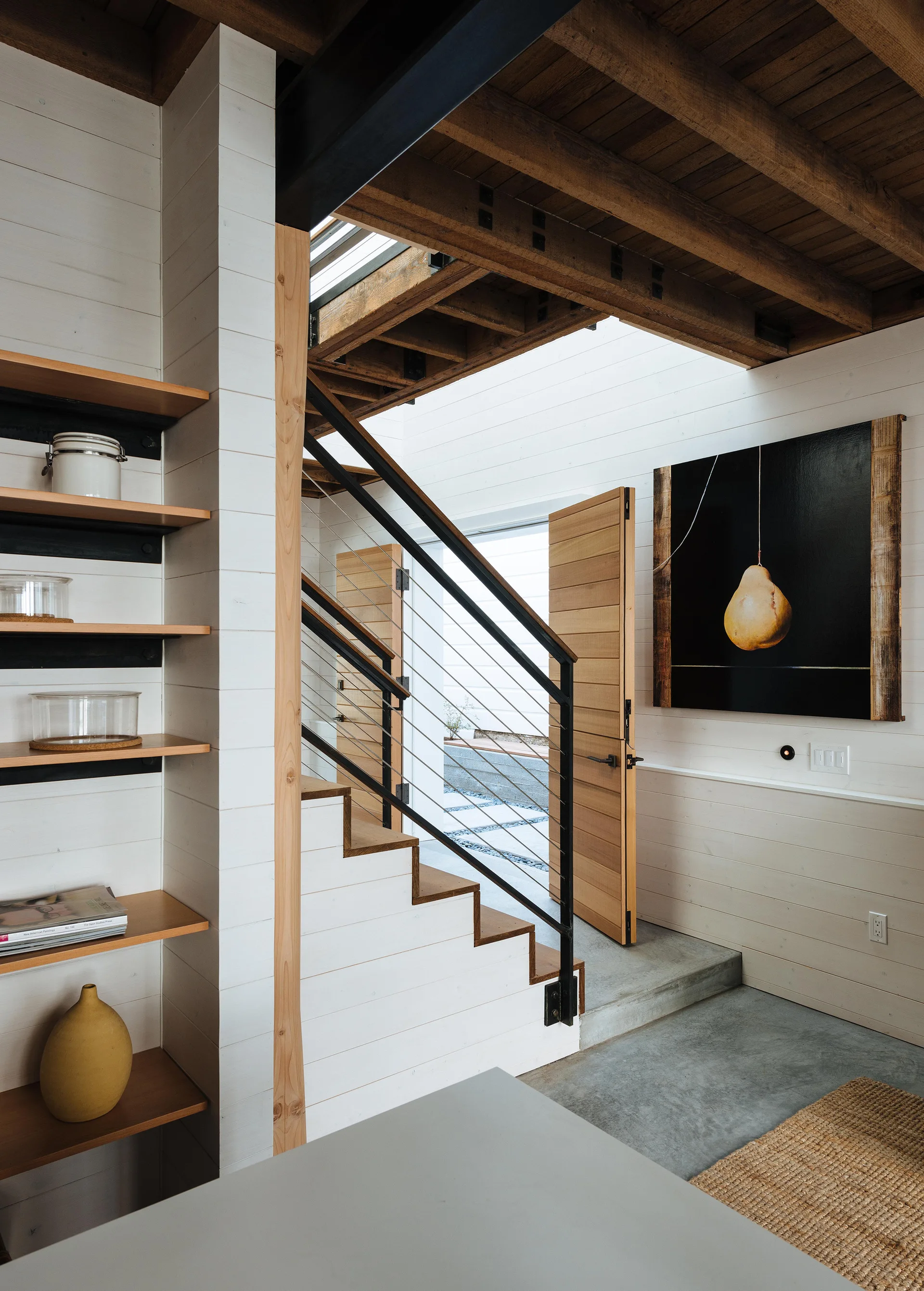 A wooden staircase with cable railings stands next to white shiplap walls, exposed timber ceiling beams, and a concrete floor.