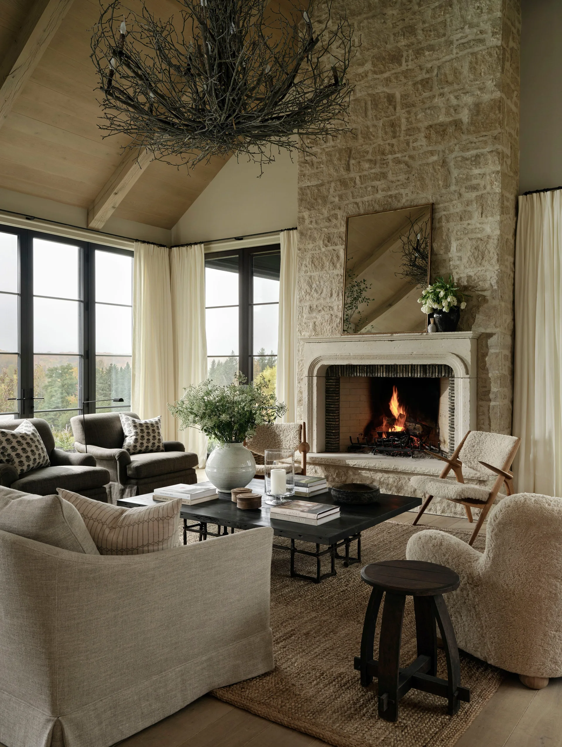 A living room with a stone fireplace, neutral armchairs around a dark coffee table, and a large branching twig chandelier under a vaulted wood ceiling.