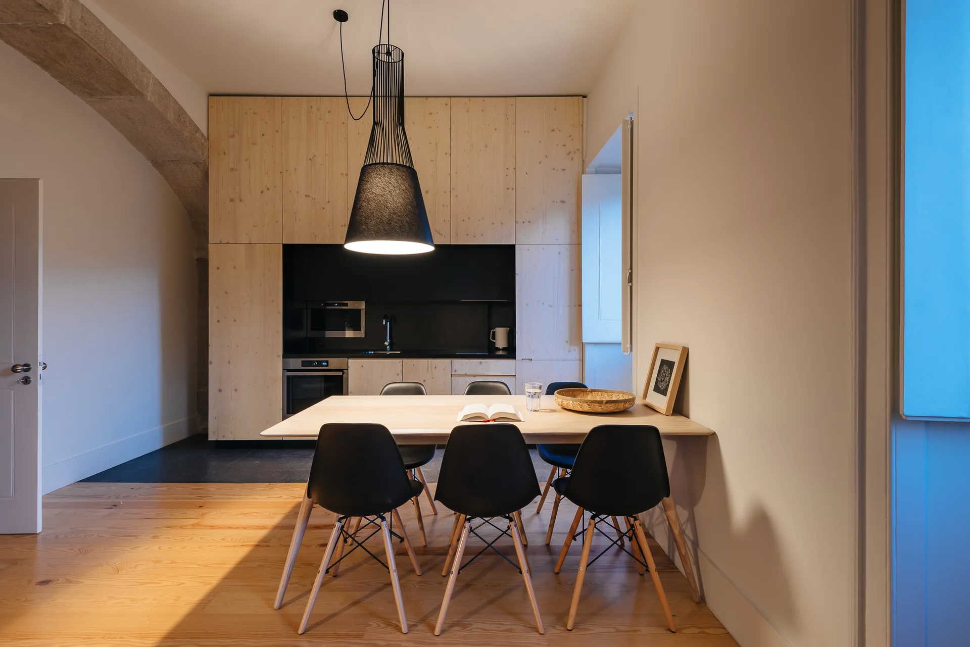 A dining room features a light wood table, black chairs, a large black pendant light, and flat-front pale wood kitchen cabinets.