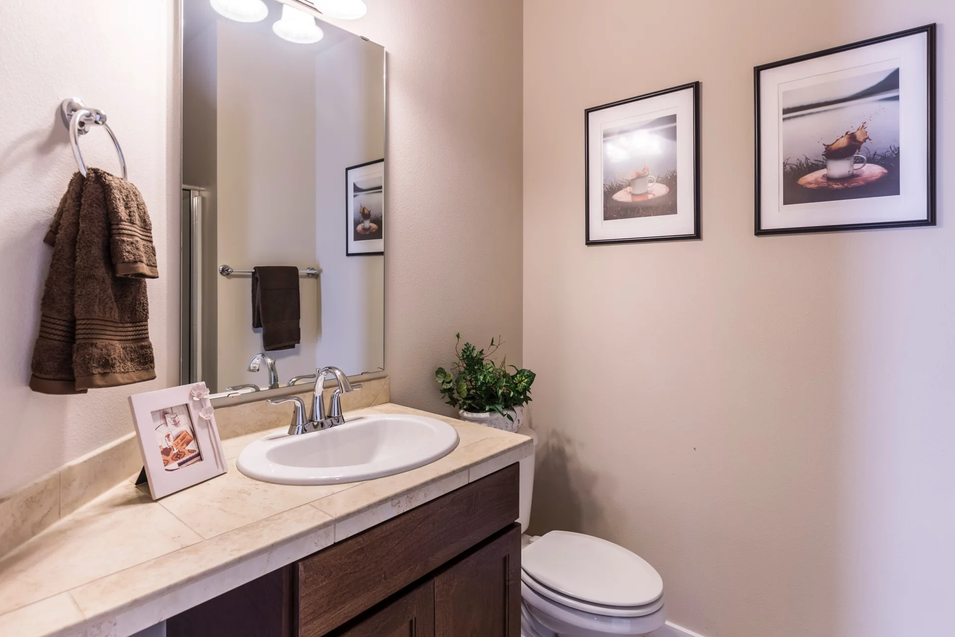 A powder room features a dark wood vanity with a beige tiled countertop, a silver faucet, and two framed prints on the beige wall.