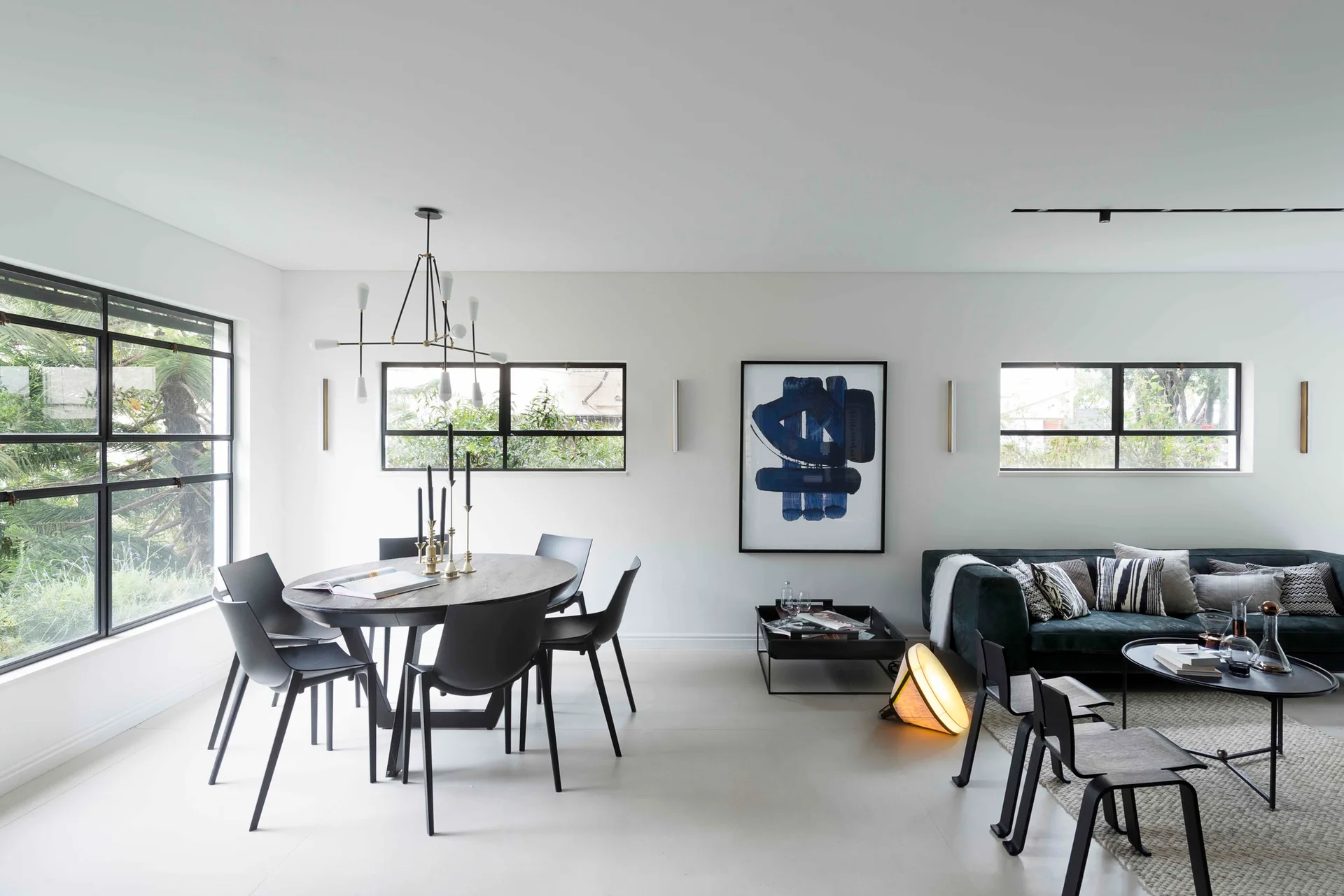 A minimalist open-plan room with a dark oval dining set, a low-profile sofa, and large black-framed windows on white walls.