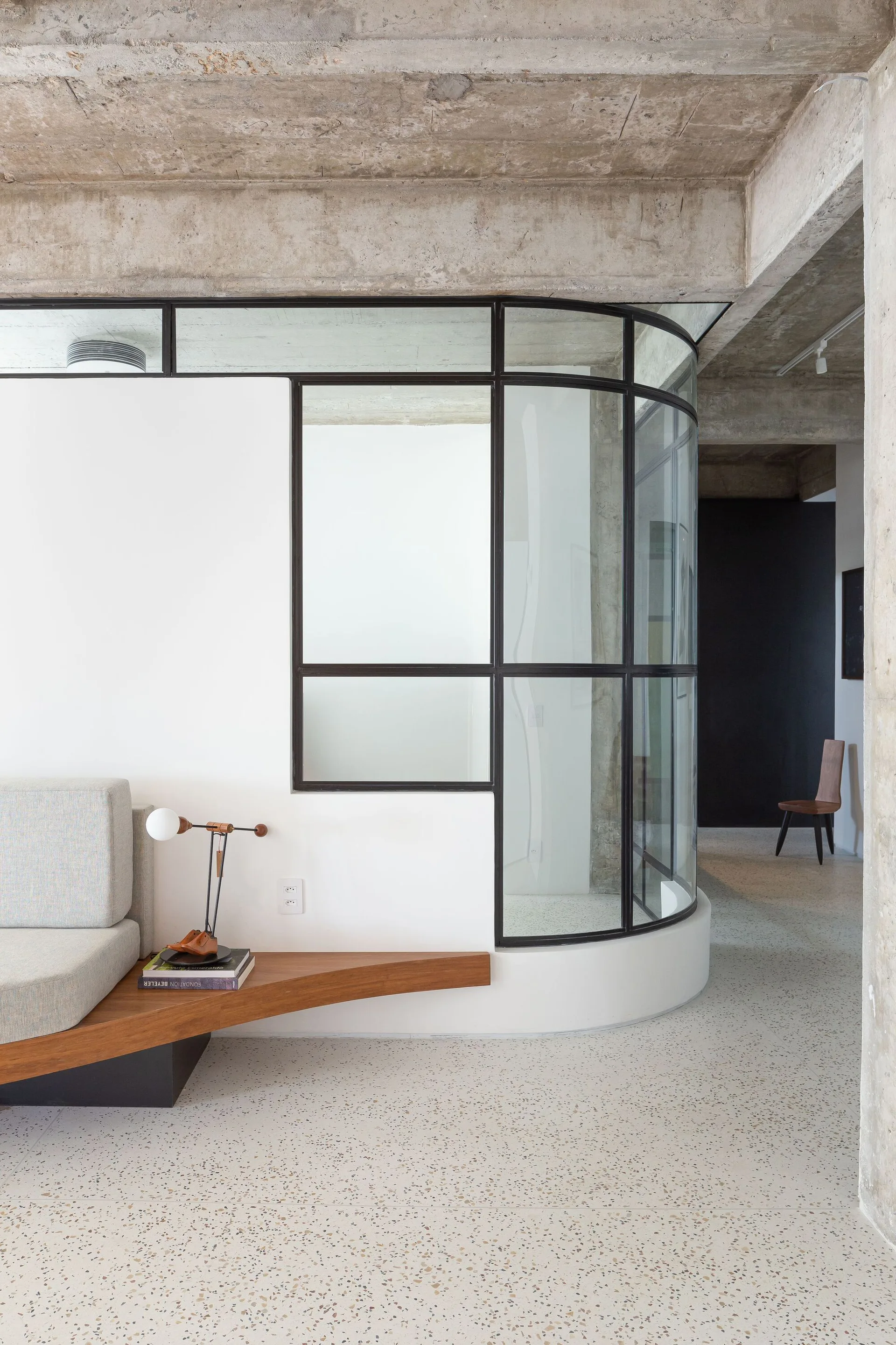 A minimalist living area features an exposed concrete ceiling, a curved glass and metal partition wall, and a low triangular wood table next to a sofa.