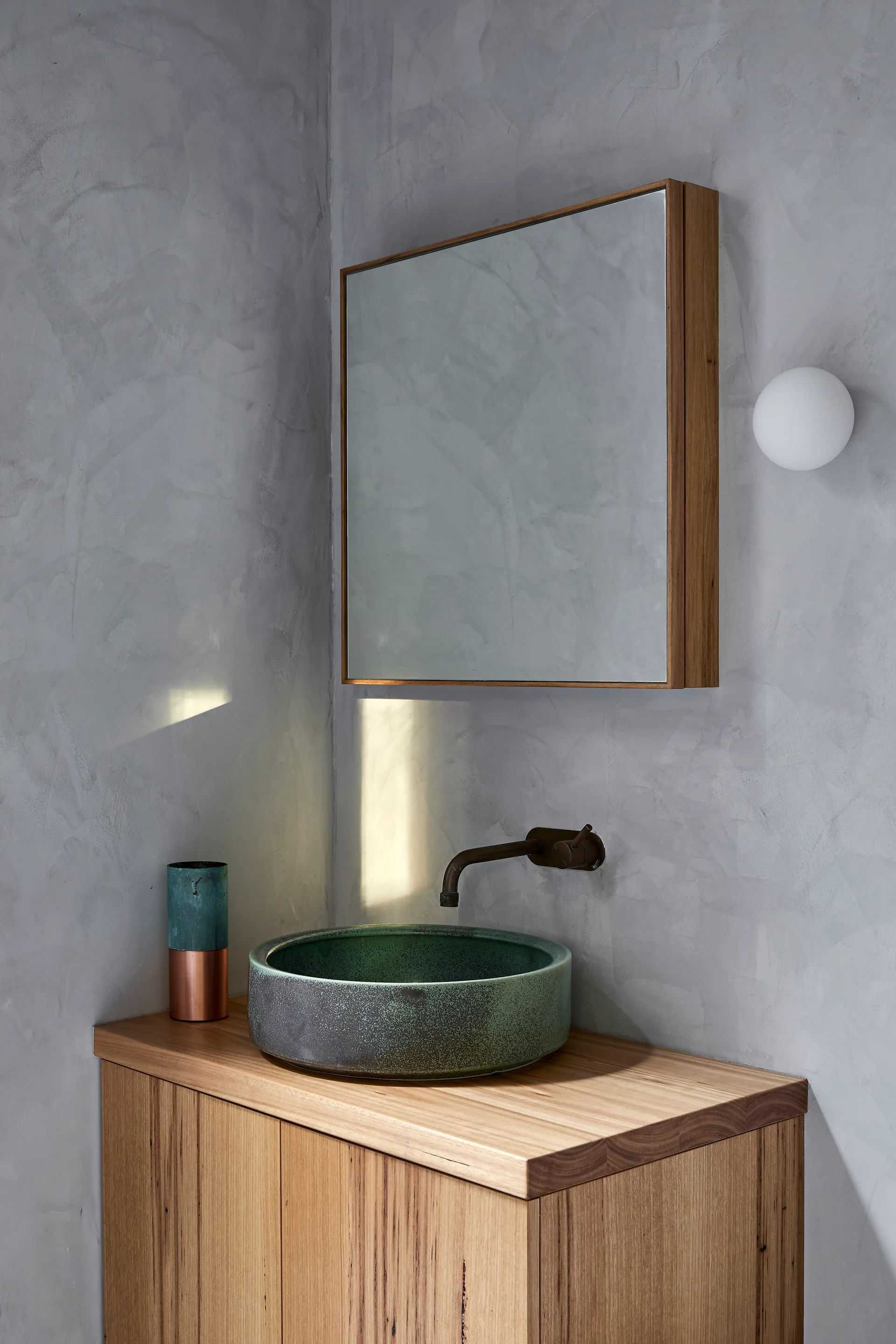A green ceramic vessel sink sits on a wood vanity cabinet against a textured gray plaster wall with a wall-mounted faucet and wood-framed mirror.