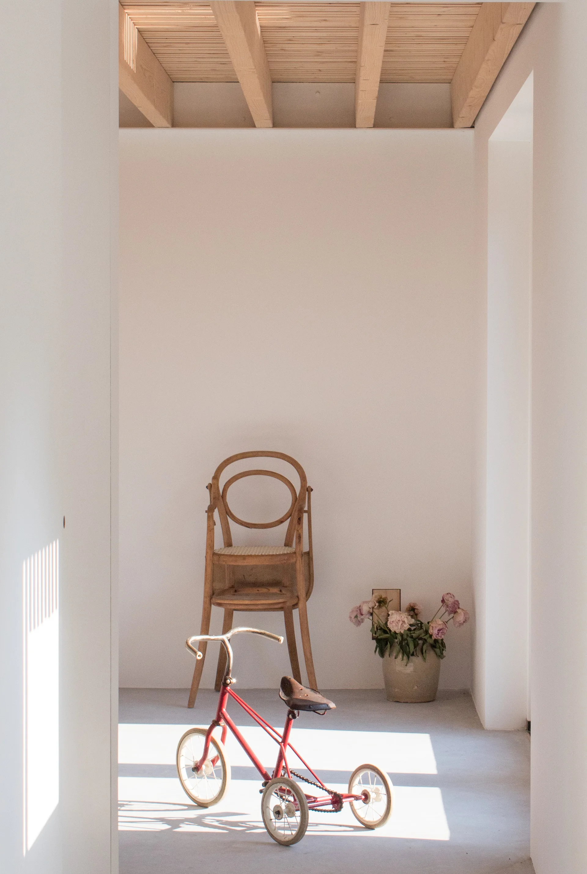 A minimalist white nook with exposed wooden ceiling beams featuring a stacked bentwood chair, a ceramic vase of flowers, and a vintage red tricycle.