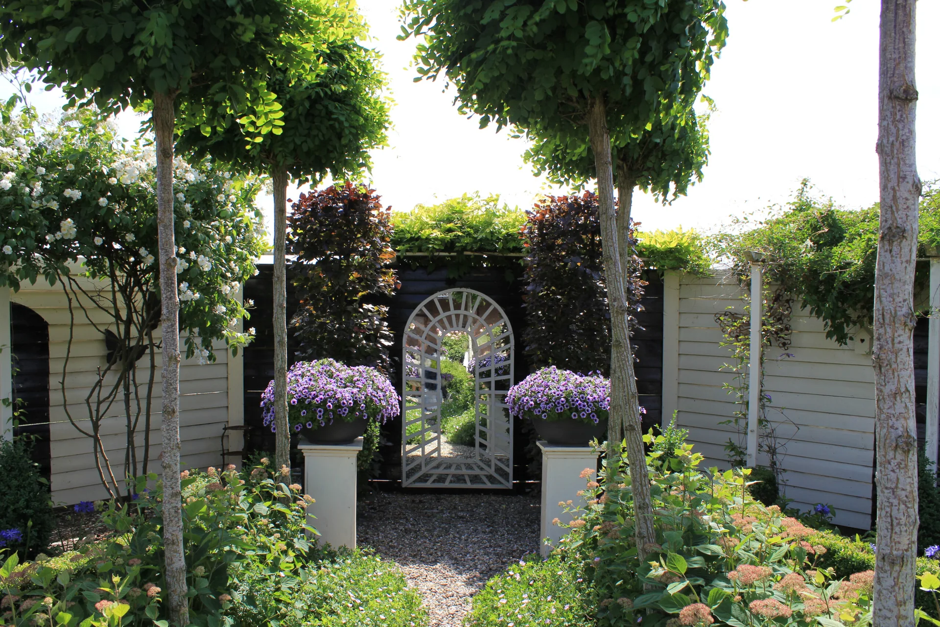 A symmetrical garden path featuring a central arched mirror on a dark wall, flanked by white pedestals with purple flowers and green topiary trees.