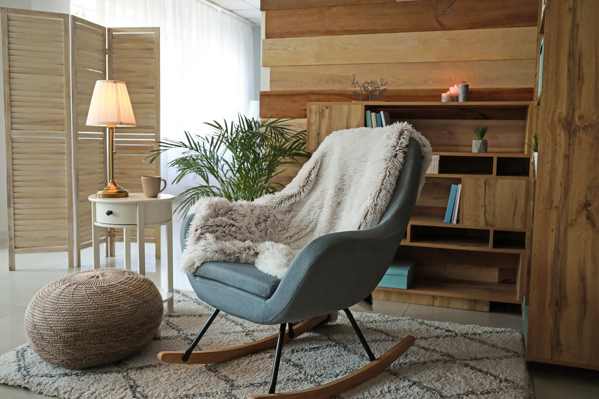 A cozy reading corner featuring an upholstered blue rocking chair with a faux fur throw blanket, a woven pouf, and a round wooden side table.