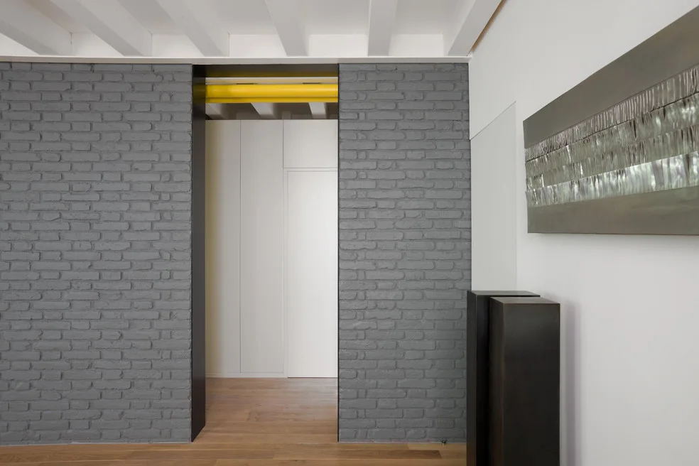 A modern transition area featuring a charcoal grey painted brick wall, exposed white ceiling beams, and a bright yellow pipe over a doorway.