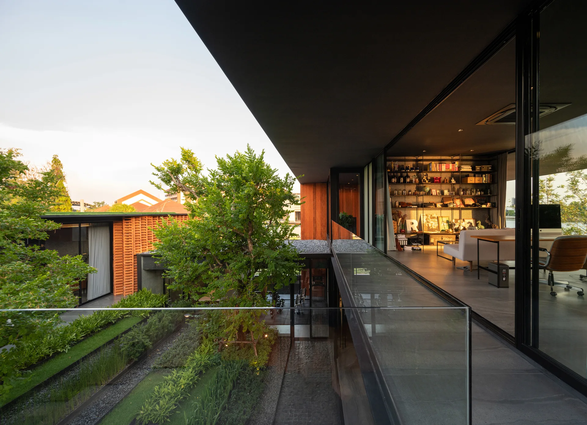 An elevated modern balcony with a glass railing looks down onto a green courtyard and into an adjacent living area with floor-to-ceiling windows.