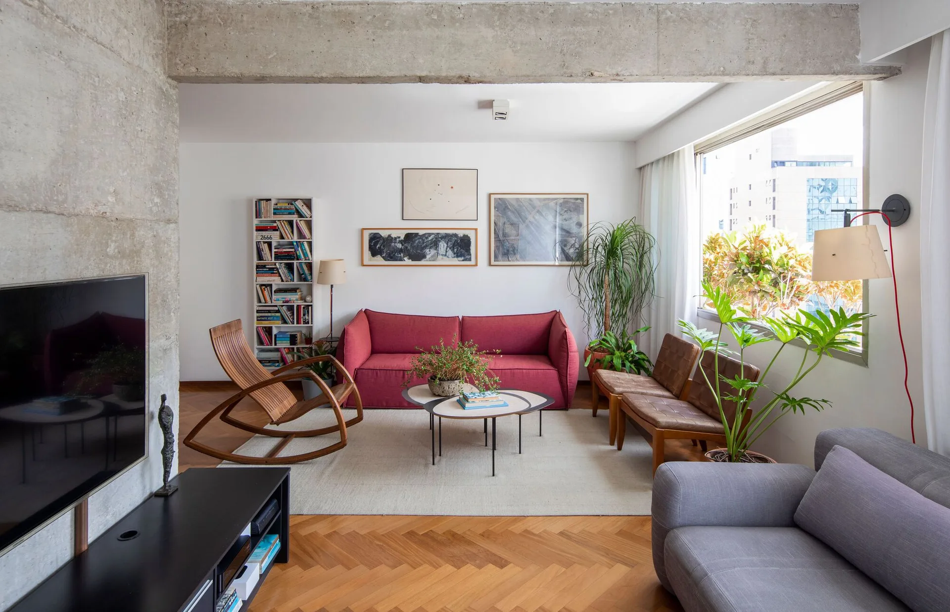 A bright living room featuring a central red sofa, mid-century wooden rocking chair, herringbone wood floors, and an exposed concrete beam.