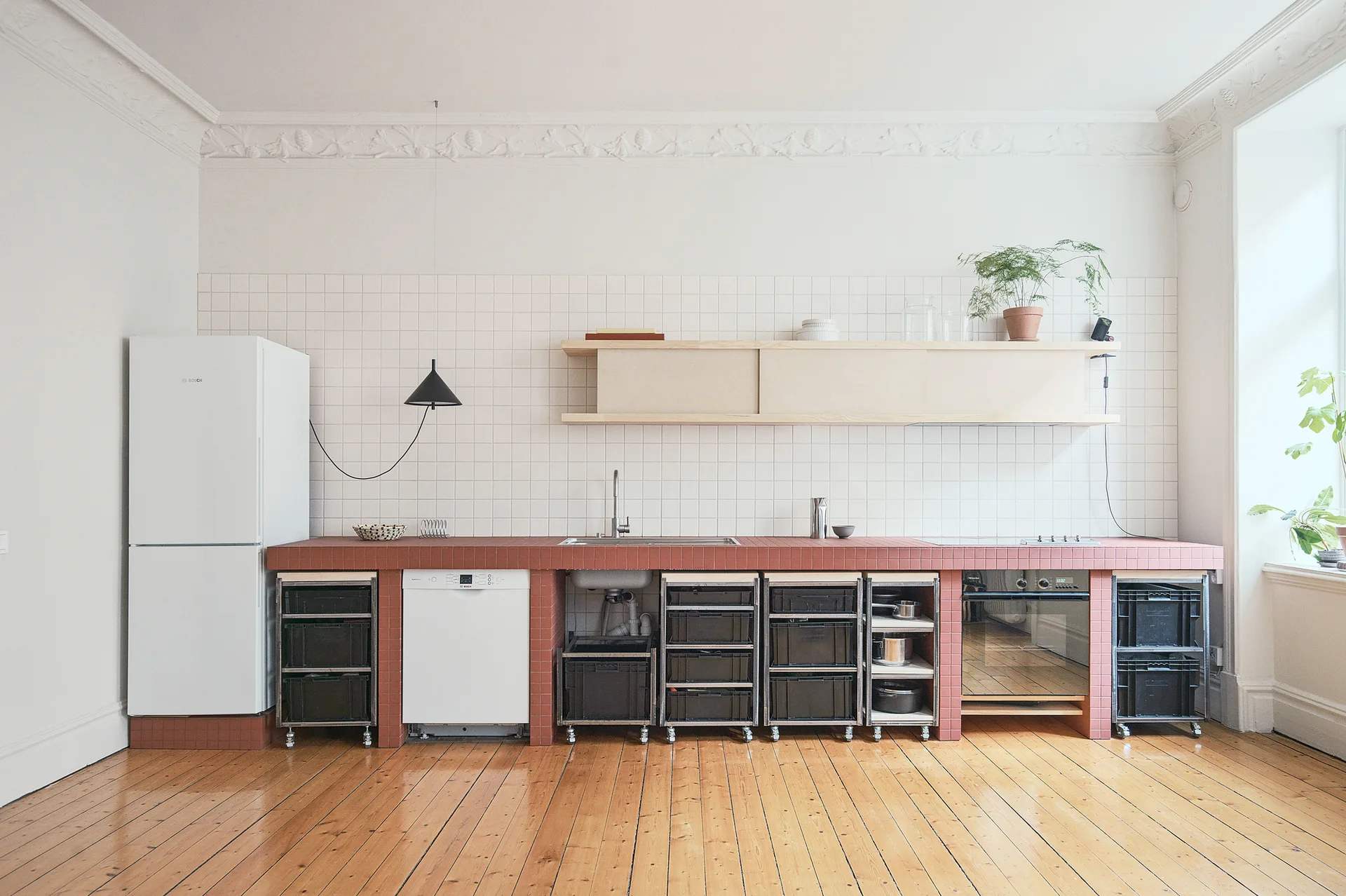 A minimalist kitchen features a long red tiled countertop, black storage crates on wheels, a white tiled backsplash, and light wood floors.