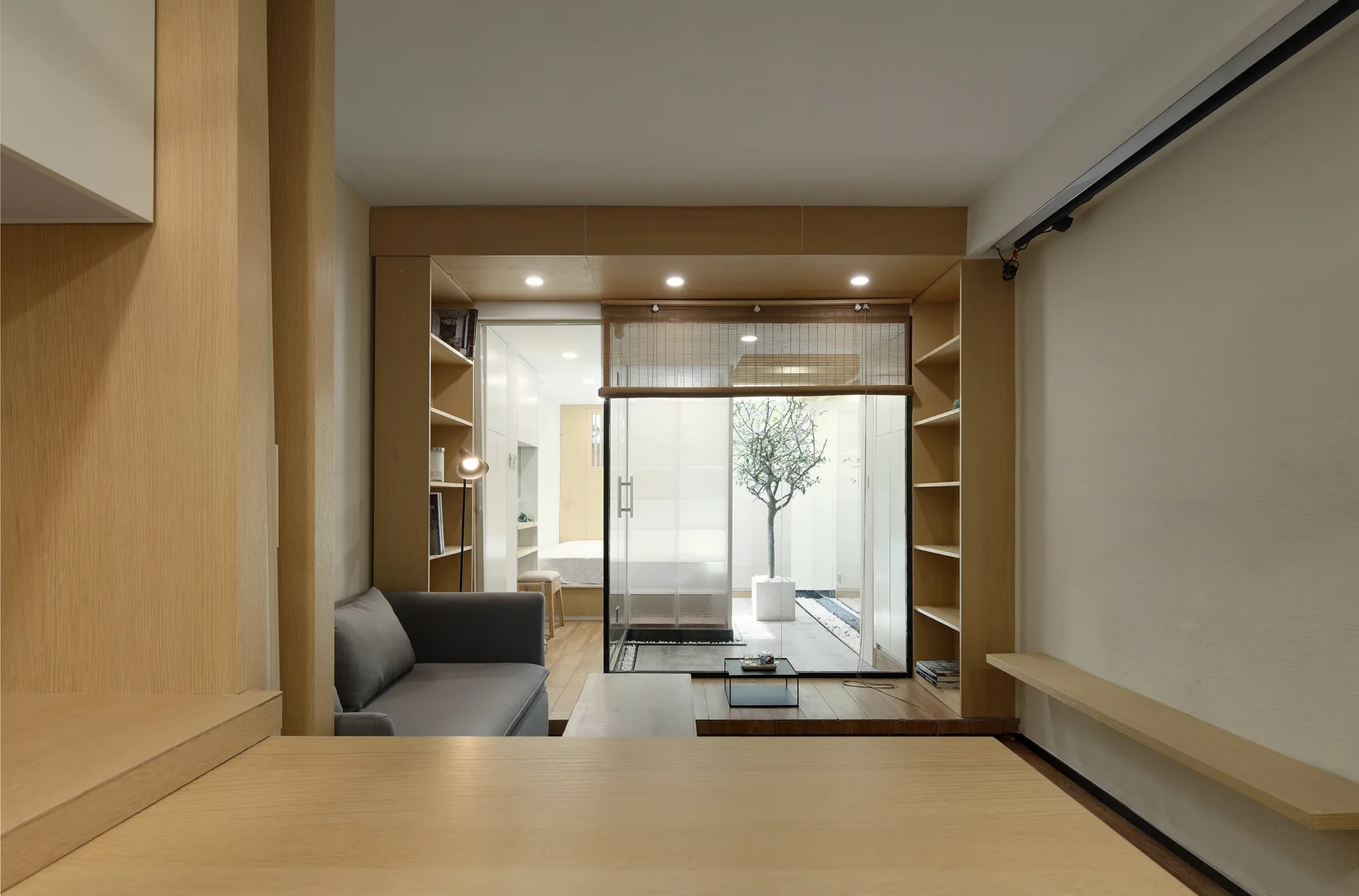 A minimalist living room features light wood built-in shelving flanking a sliding glass door that opens to a small interior courtyard.