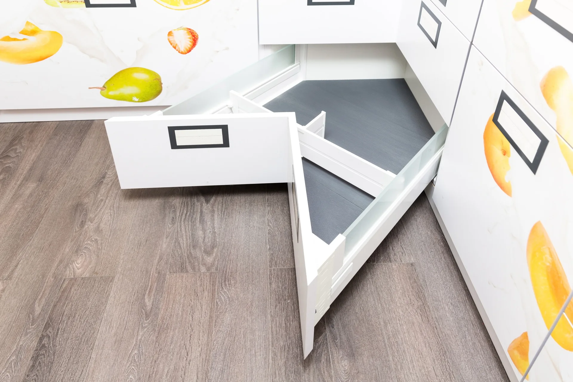 An open white corner kitchen drawer with a grey interior and dividers sits above wood flooring.