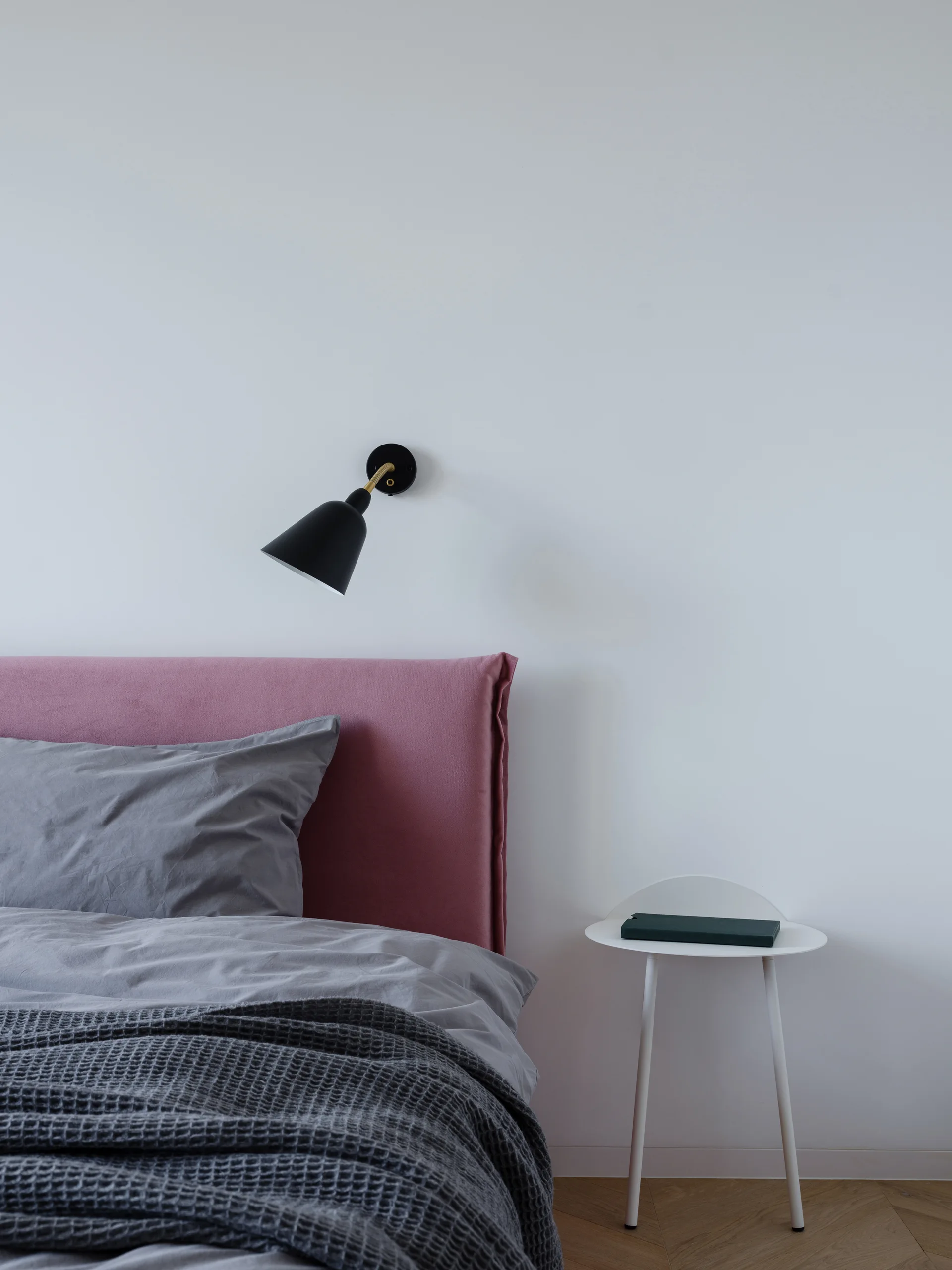 A pink upholstered headboard with grey bedding next to a small white side table and a wall-mounted black sconce.