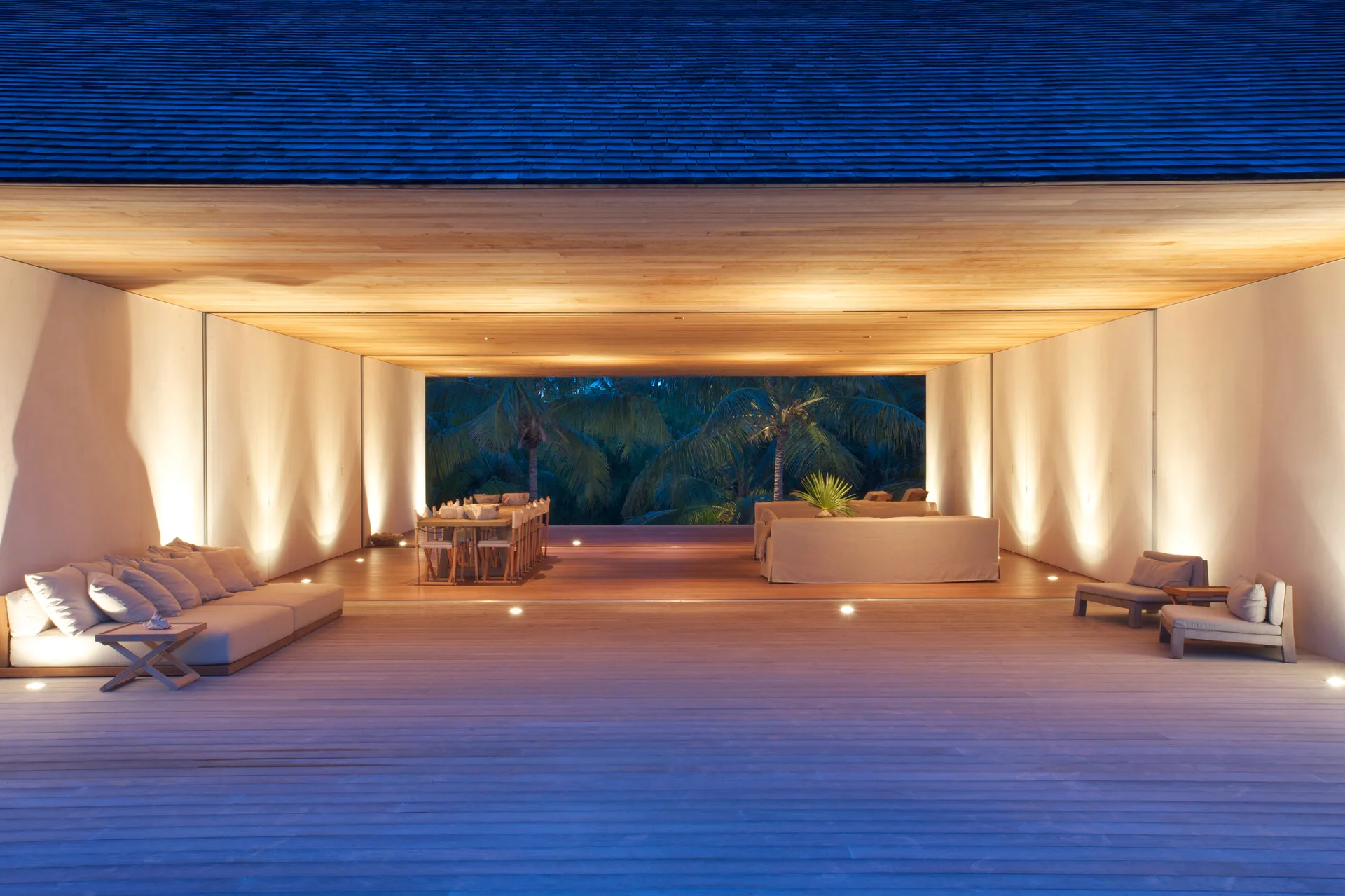 A spacious open-air wooden pavilion features low-profile beige lounge sofas, a long dining table, and walls illuminated by warm uplights at dusk.
