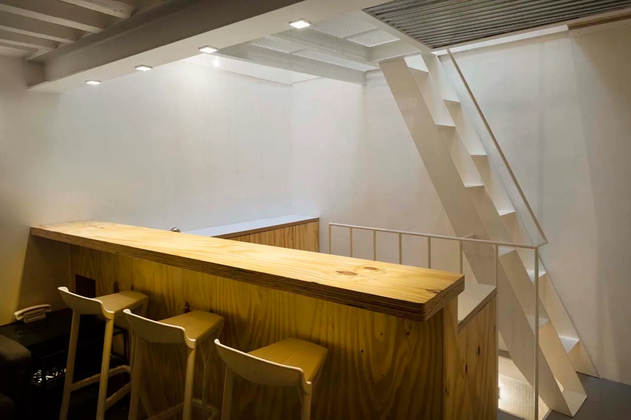 A modern interior features a plywood bar counter with three wooden stools next to a minimalist white staircase leading to an upper level.