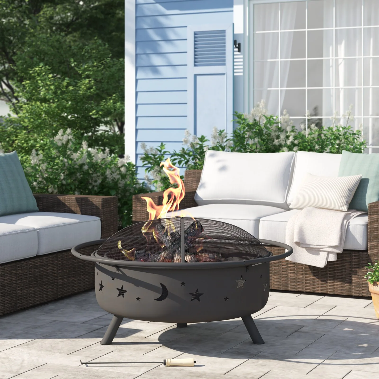 A grey stone patio features a black metal fire pit with celestial cutouts positioned in front of wicker outdoor seating with white cushions.