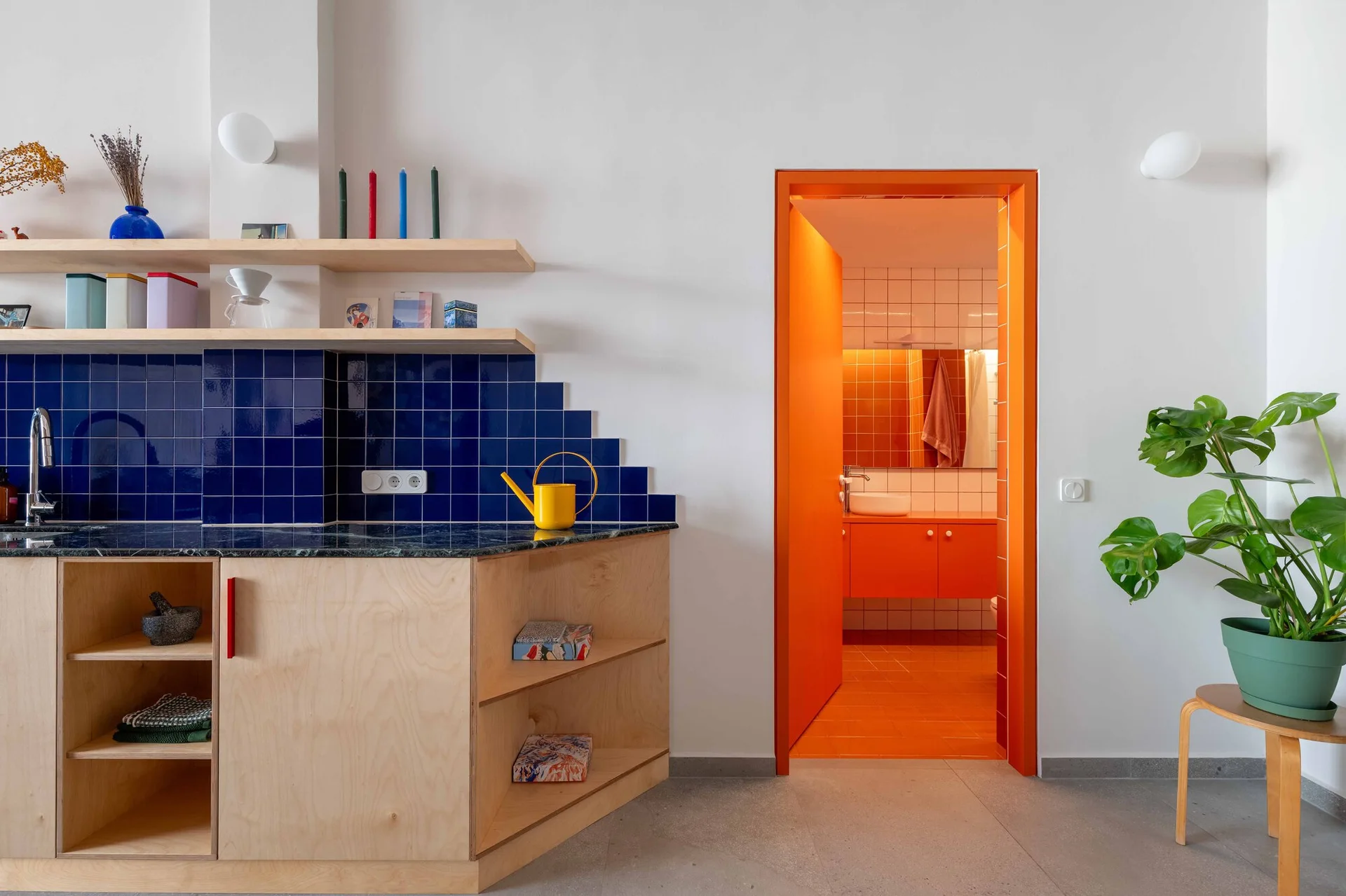 A modern kitchen featuring light wood cabinets, a cobalt blue tile backsplash, and a vibrant orange doorway opening to a matching orange bathroom.