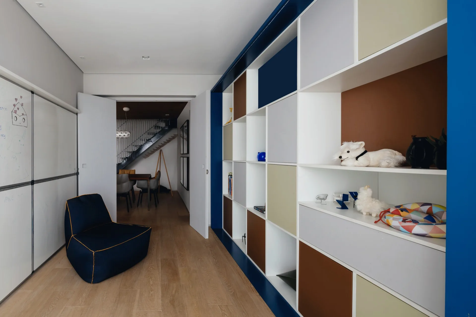 A light-filled playroom features a large, colorful built-in grid shelving unit, a dark beanbag chair, and a whiteboard wall.