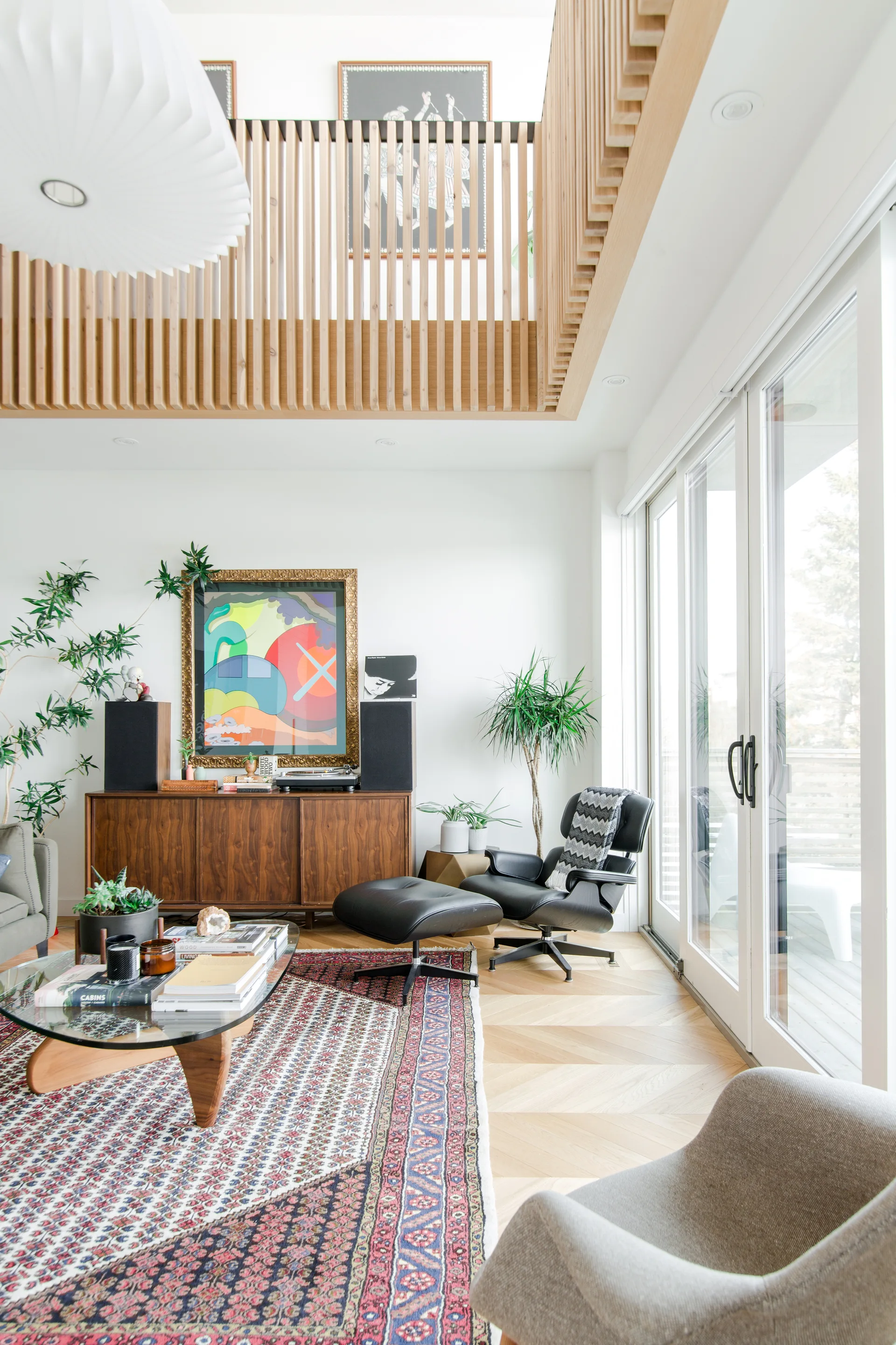 A bright mid-century modern living room featuring a black leather lounge chair, a walnut sideboard, herringbone wood flooring, and a slatted wood balcony.
