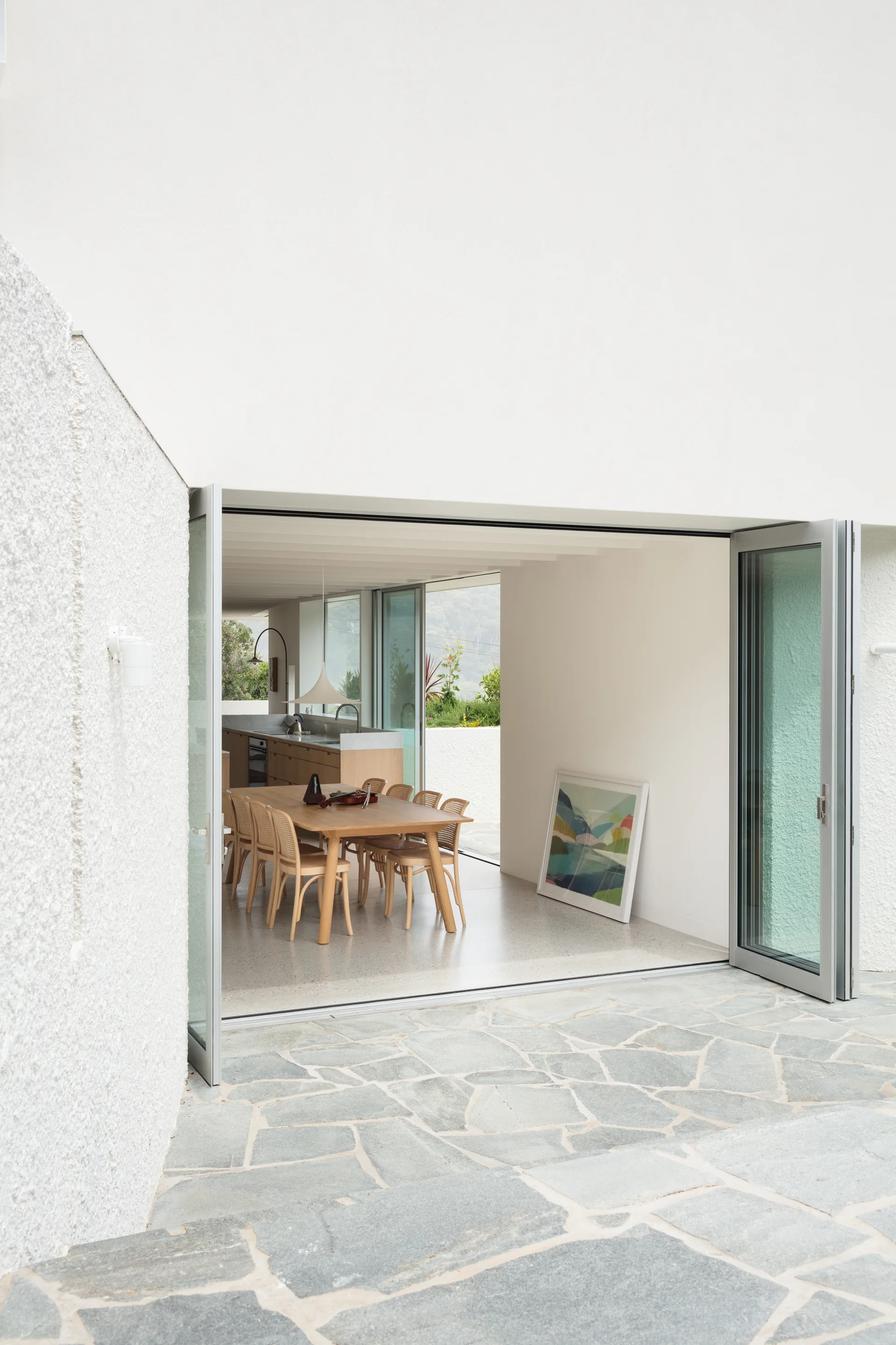 A view from a grey flagstone patio through open bifold glass doors into a white dining room with a wooden table and cane chairs.