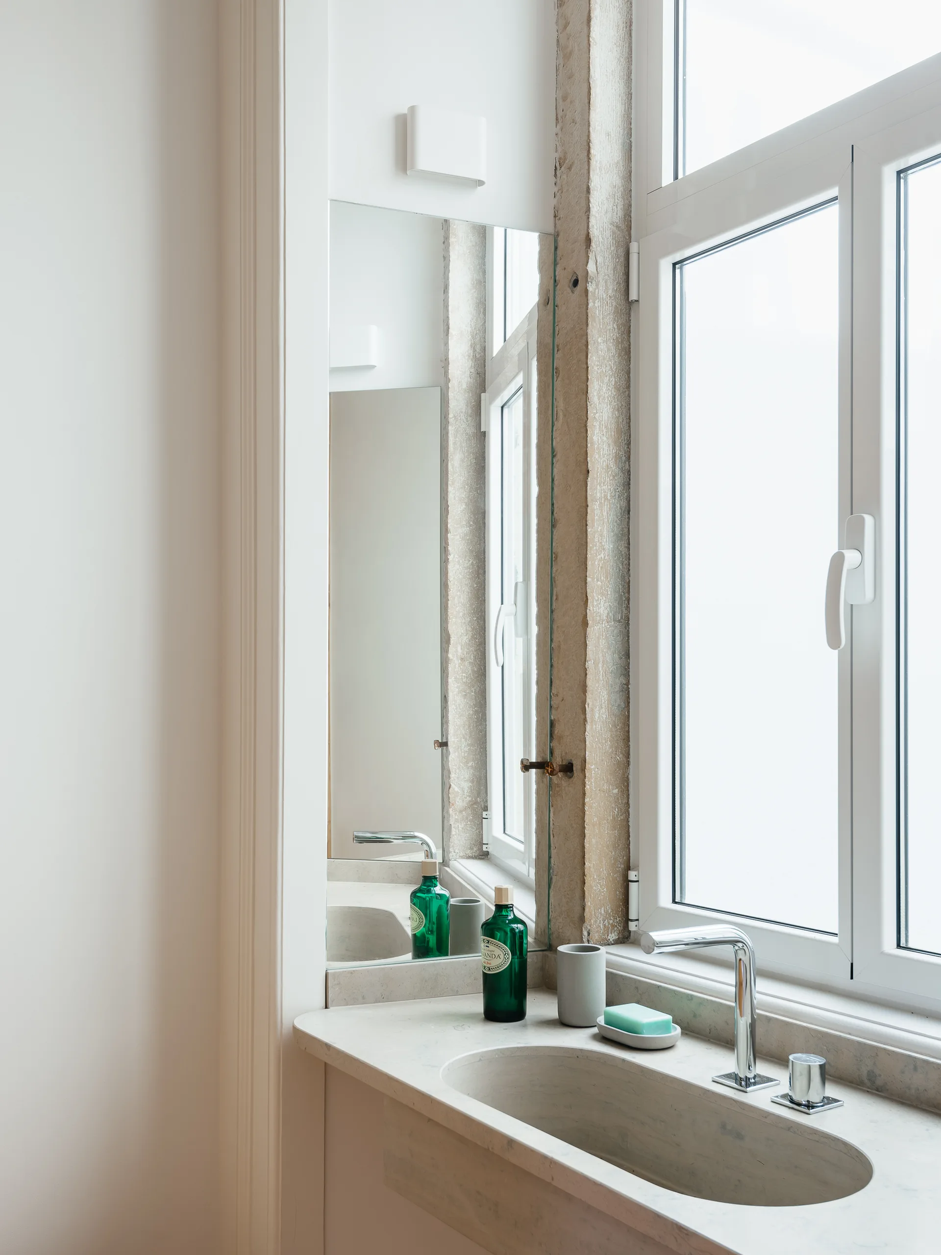 A minimalist bathroom vanity featuring an integrated stone sink, a chrome faucet, a tall mirror, and an adjacent white casement window.