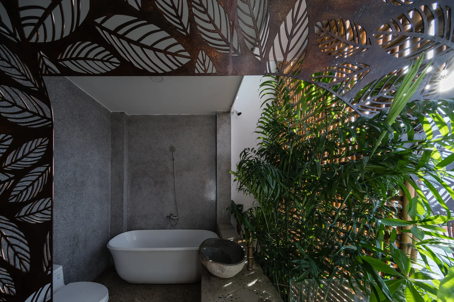 A modern bathroom features a white freestanding tub, a stone vessel sink, indoor palms, and a decorative leaf-patterned metal screen.