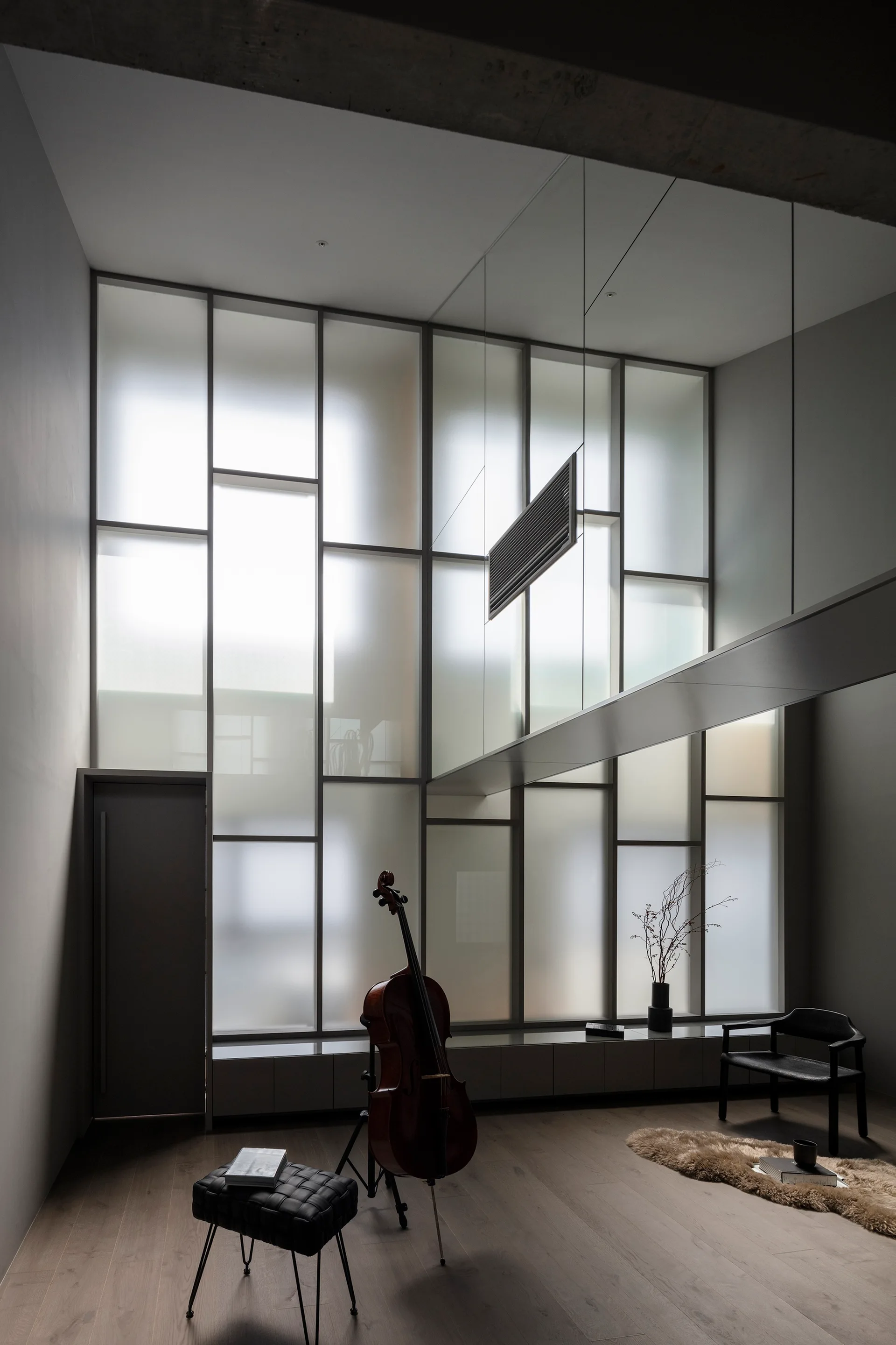 A minimalist grey living room featuring a tall frosted glass window wall, a central cello on a stand, and a black tufted leather ottoman.