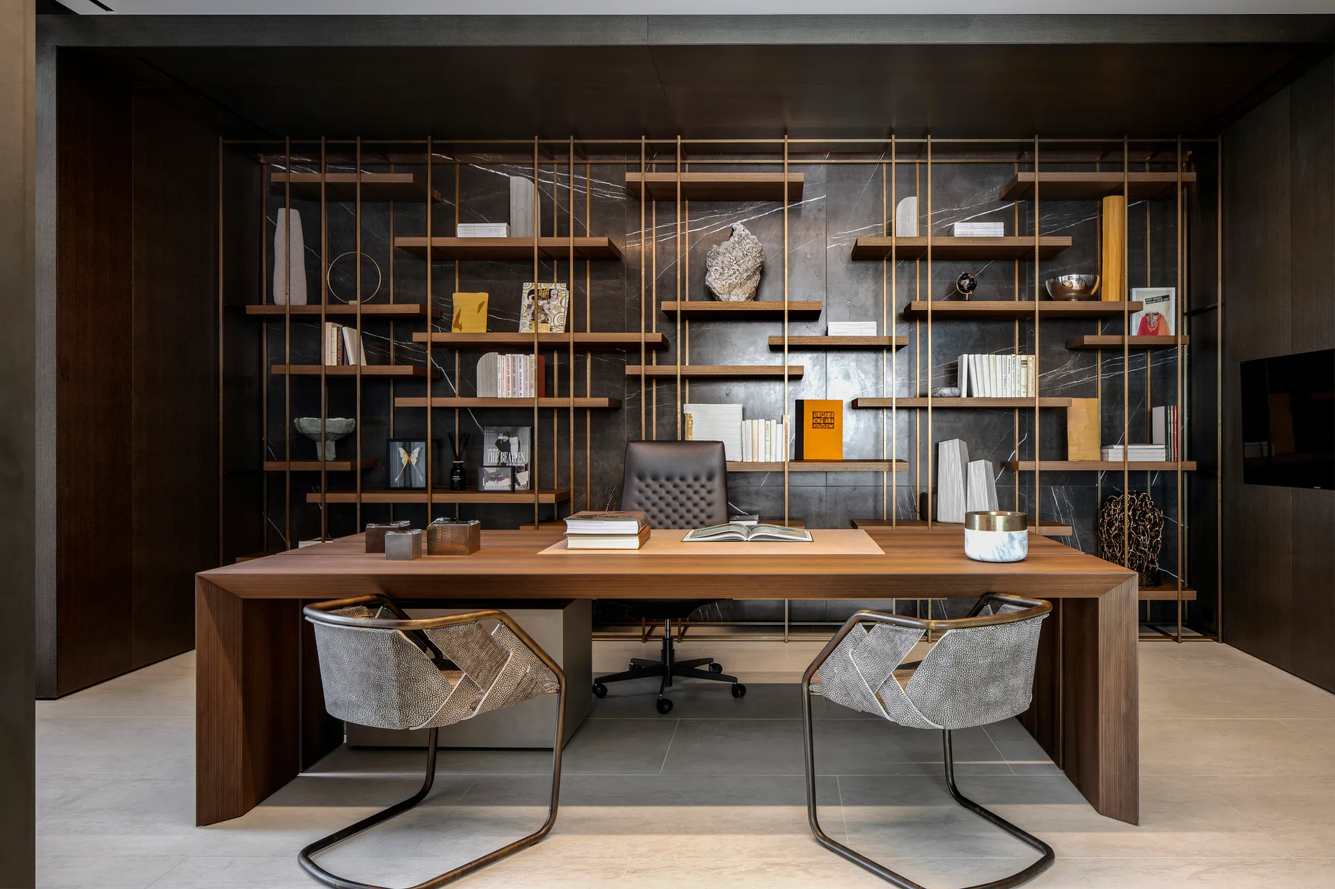 A modern home office featuring a large wooden desk, two patterned armchairs, and custom shelving mounted on a dark veined stone accent wall.