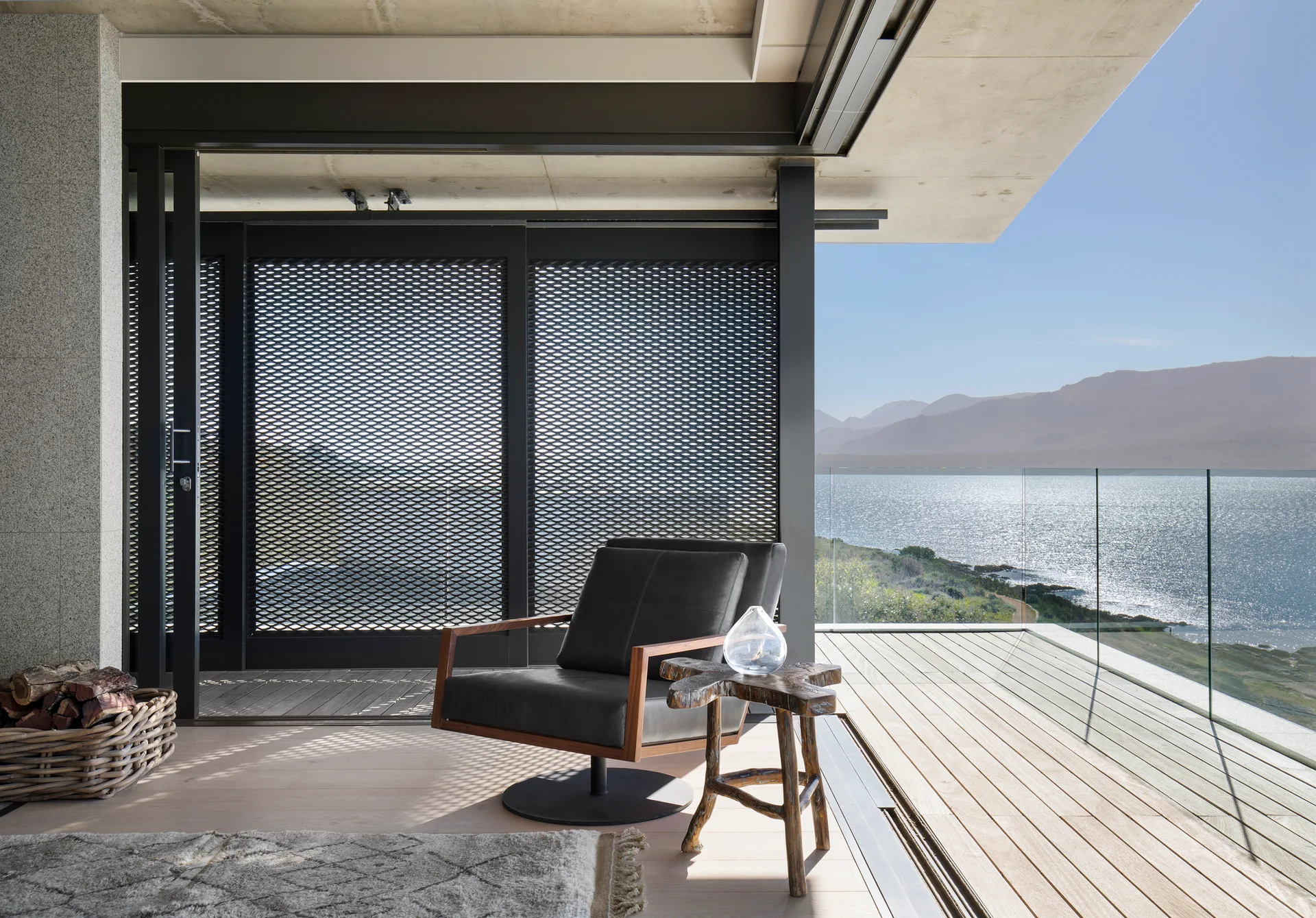 A modern balcony deck features a leather armchair and a wood stump table looking out over a lake and distant mountains behind a glass railing.