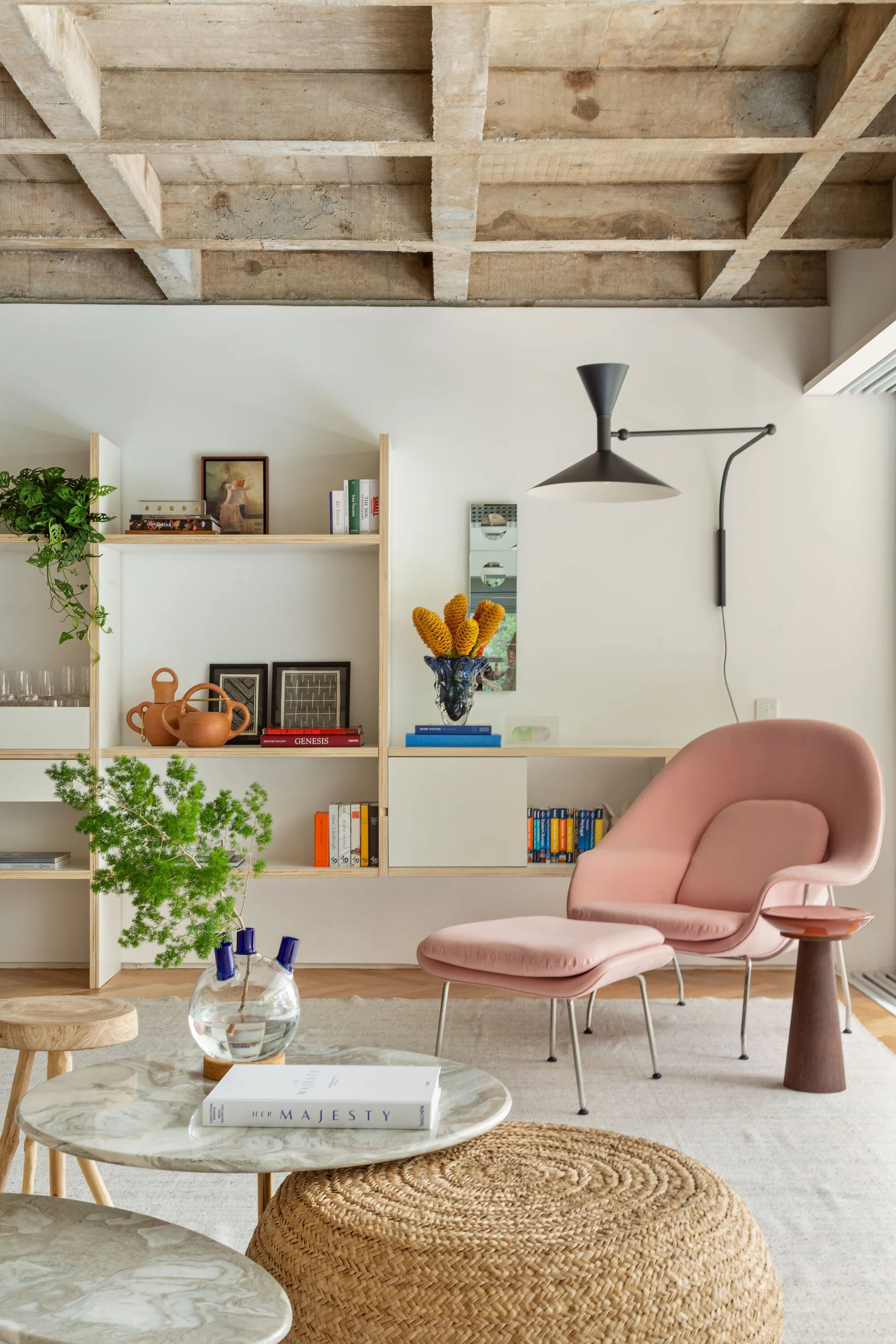 A modern living room features a pink lounge chair and ottoman, built-in wooden shelving, and an exposed concrete waffle ceiling.