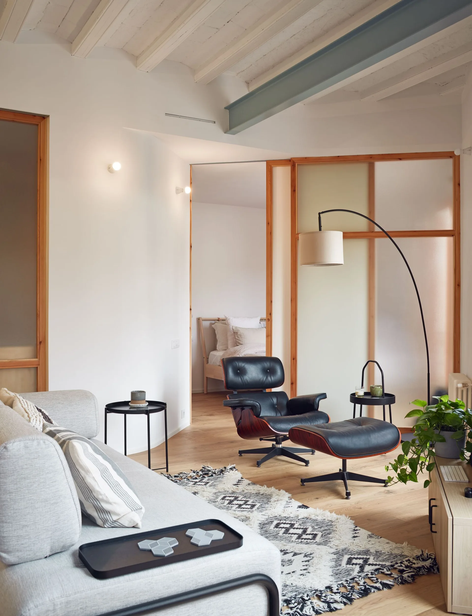 A living room featuring a grey sofa, a black leather lounge chair and ottoman, and a frosted glass partition wall leading to a bedroom.
