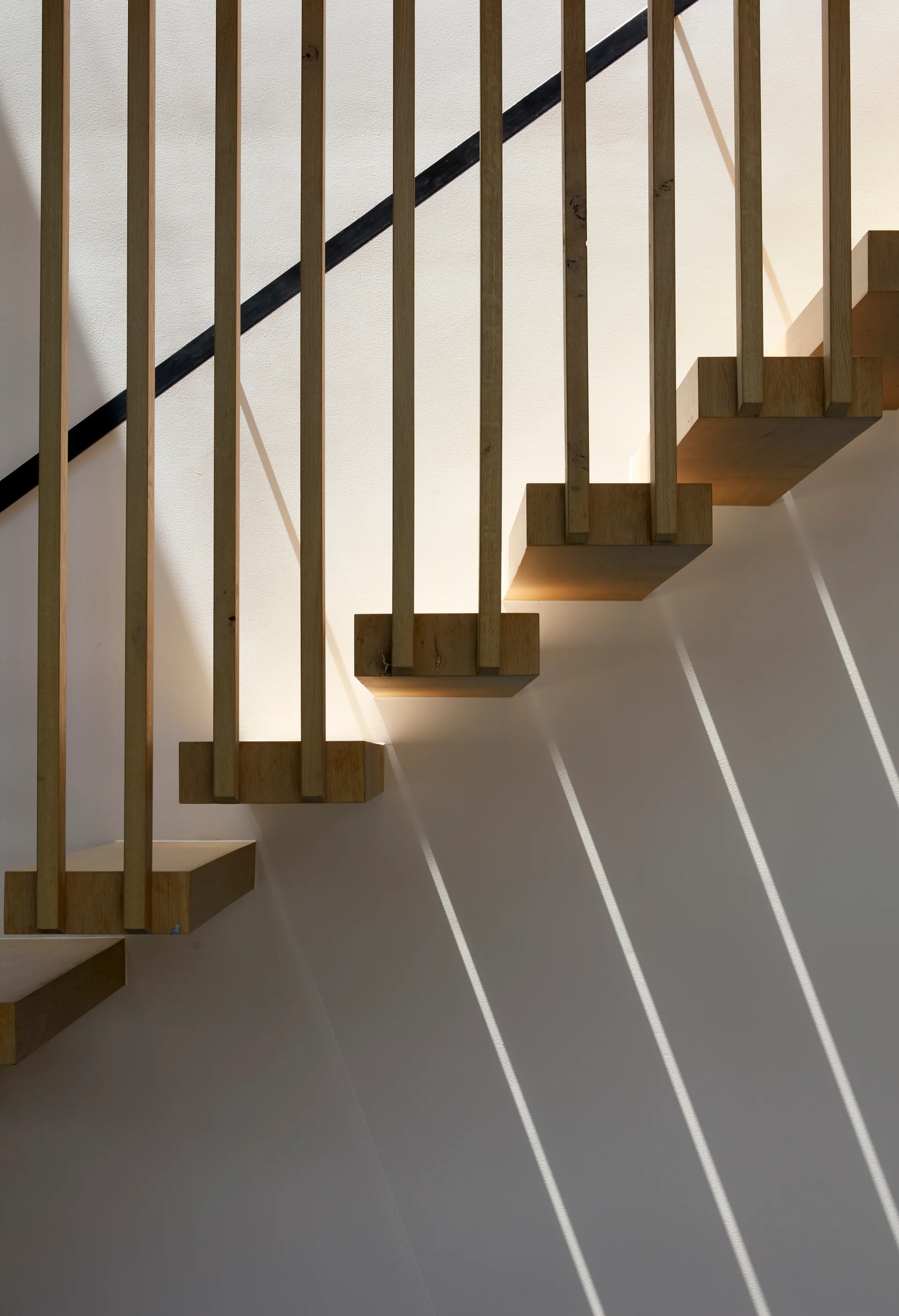 Floating light wood stair treads are supported by vertical wooden slats on a white wall, casting geometric shadows under direct light.