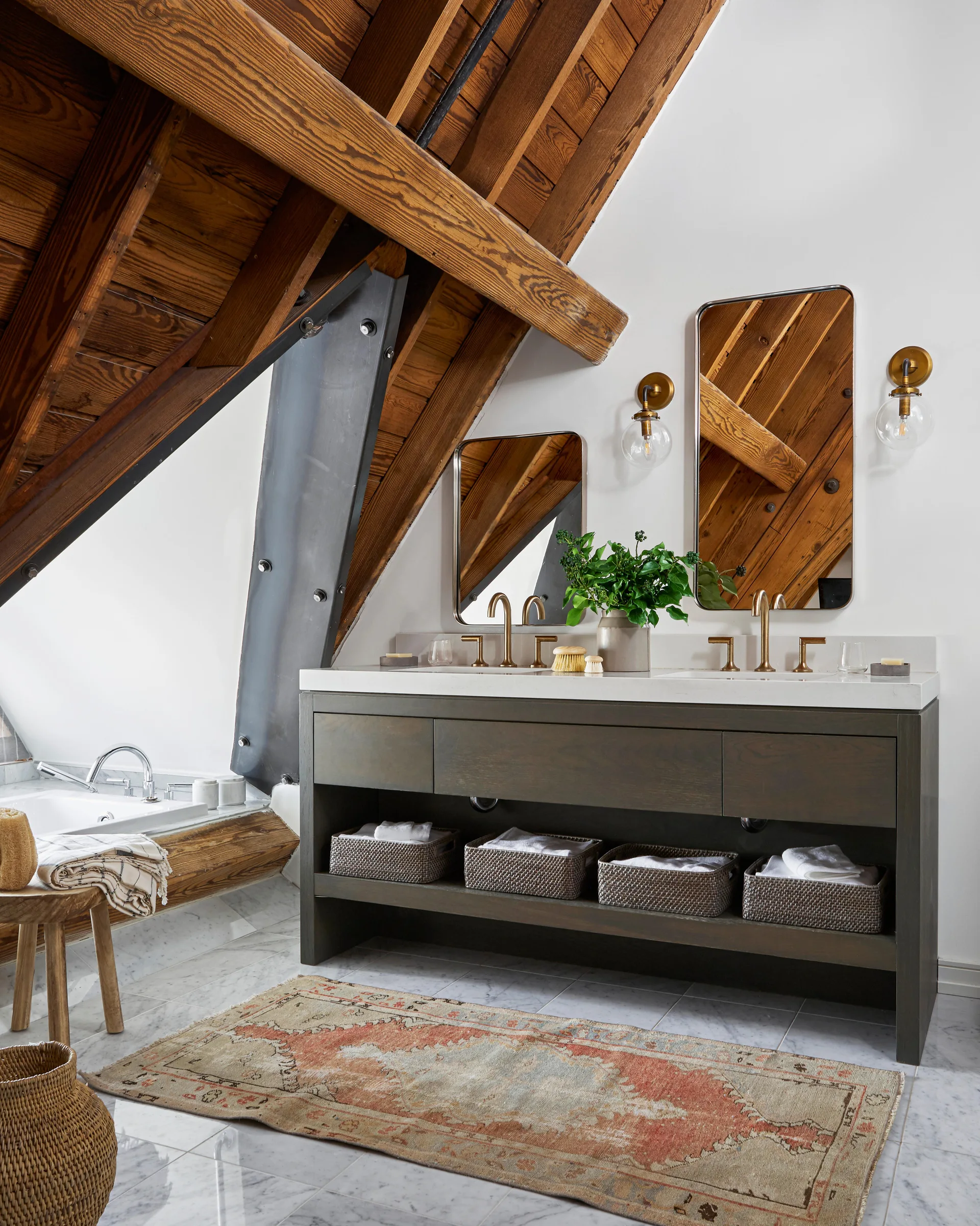 An attic bathroom features a dark double vanity with gold fixtures beneath a sloped wooden ceiling with exposed rafters.